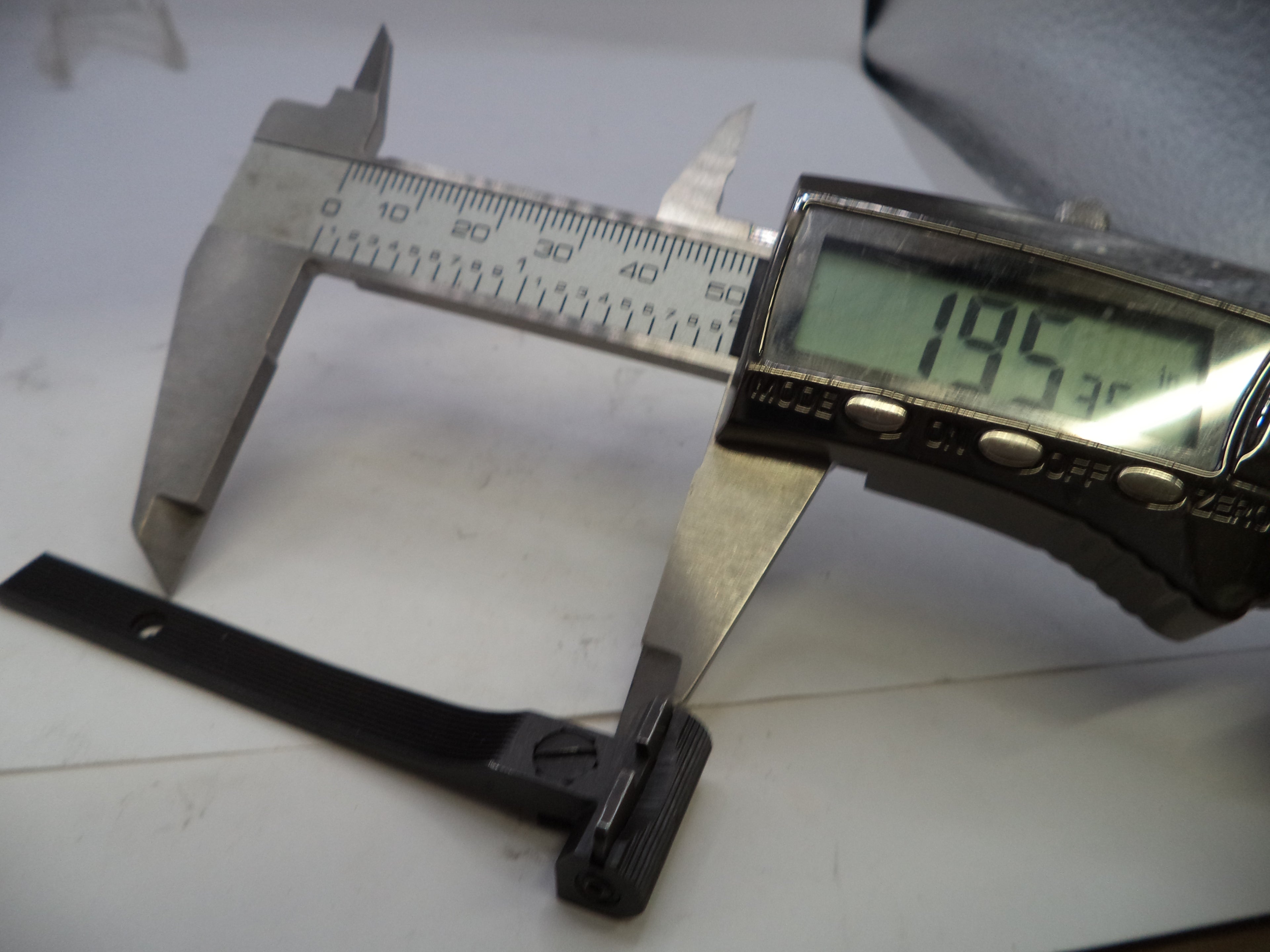 Digital caliper measuring a black metal bracket on a white surface
