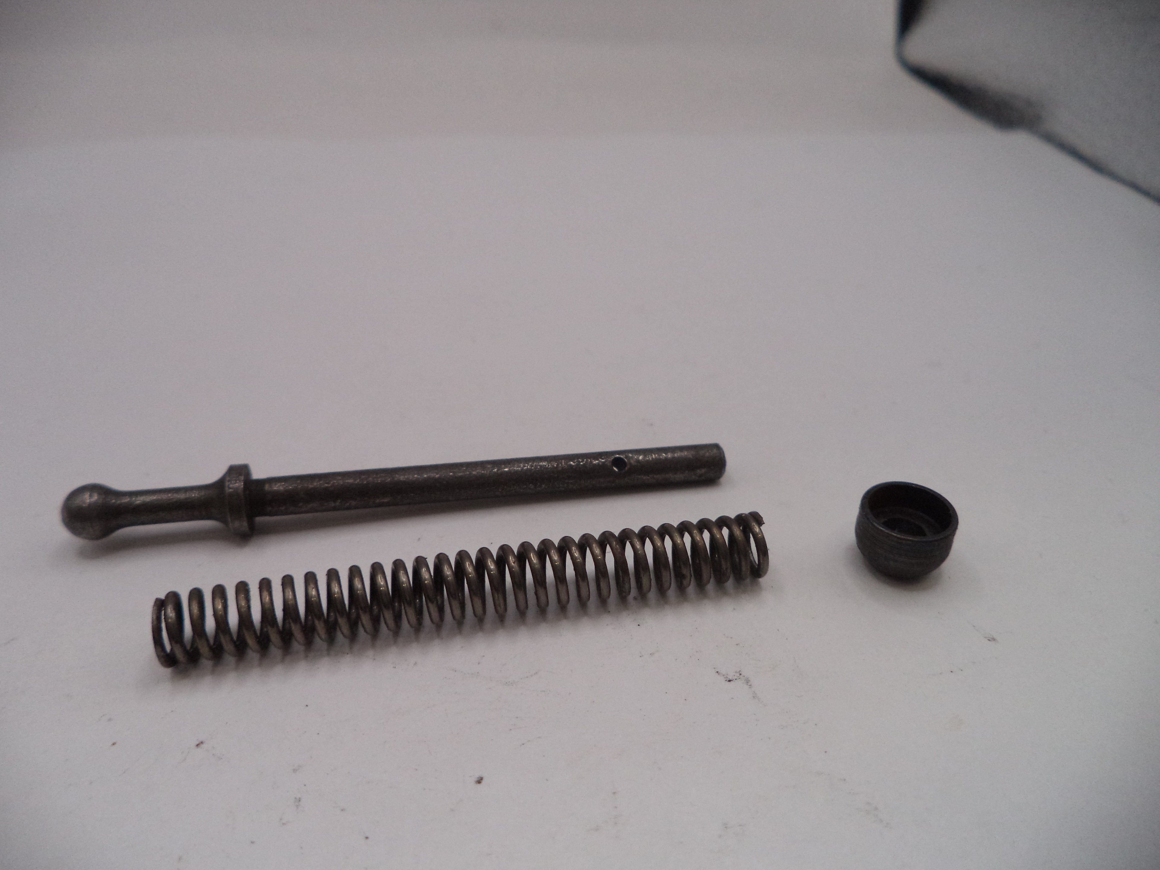 Mechanical components including a spring, rod, and small black cap on a white surface.