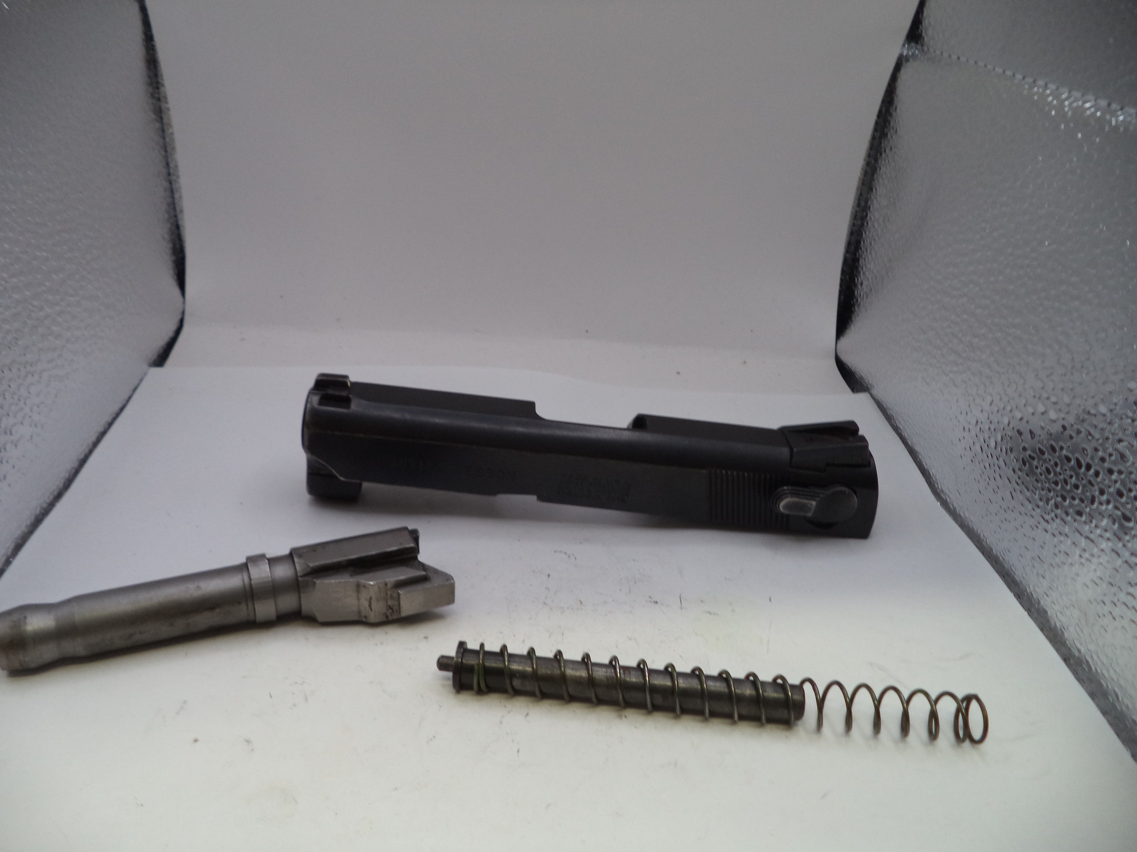 Gun components including a barrel, slide, and spring on a white surface with a gray background.