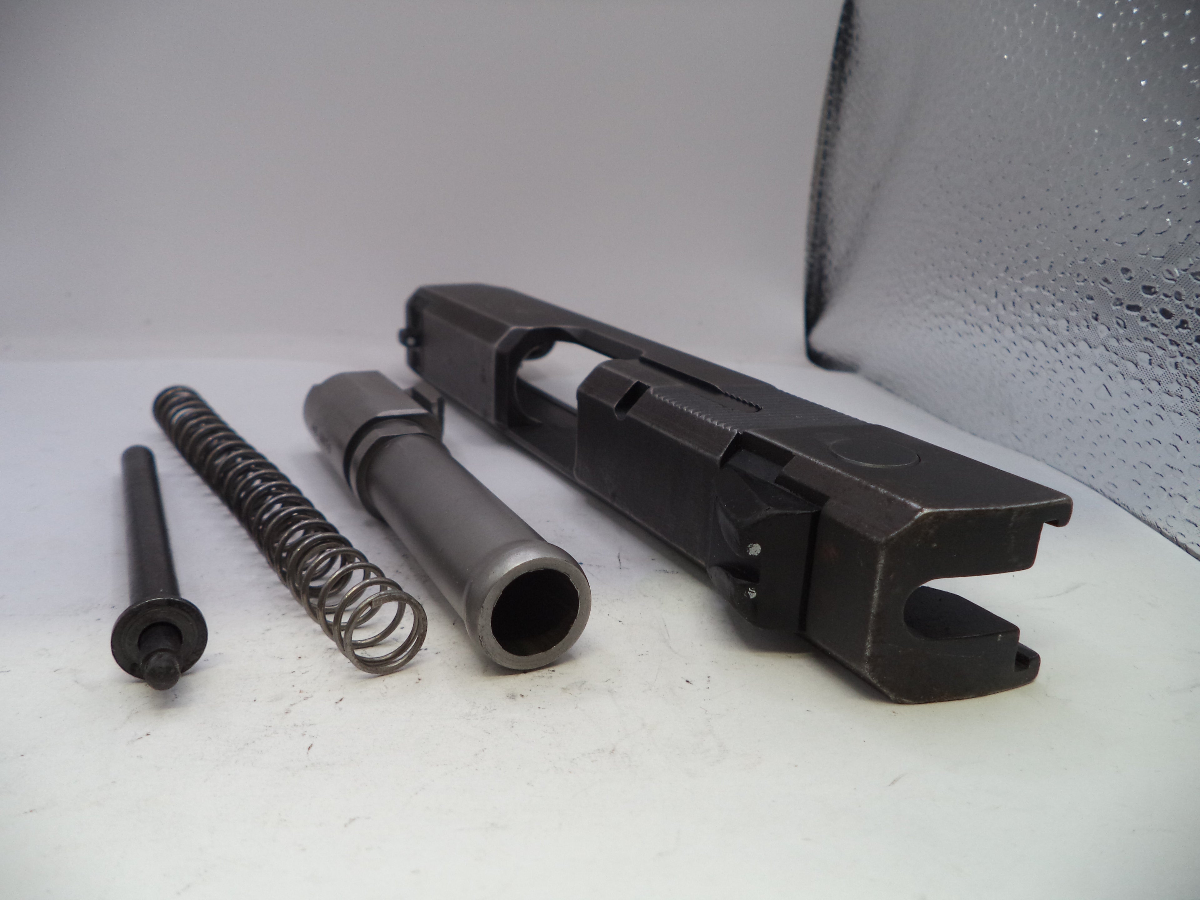 Black metal gun slide, spring, and other components on a white surface with a gray background.
