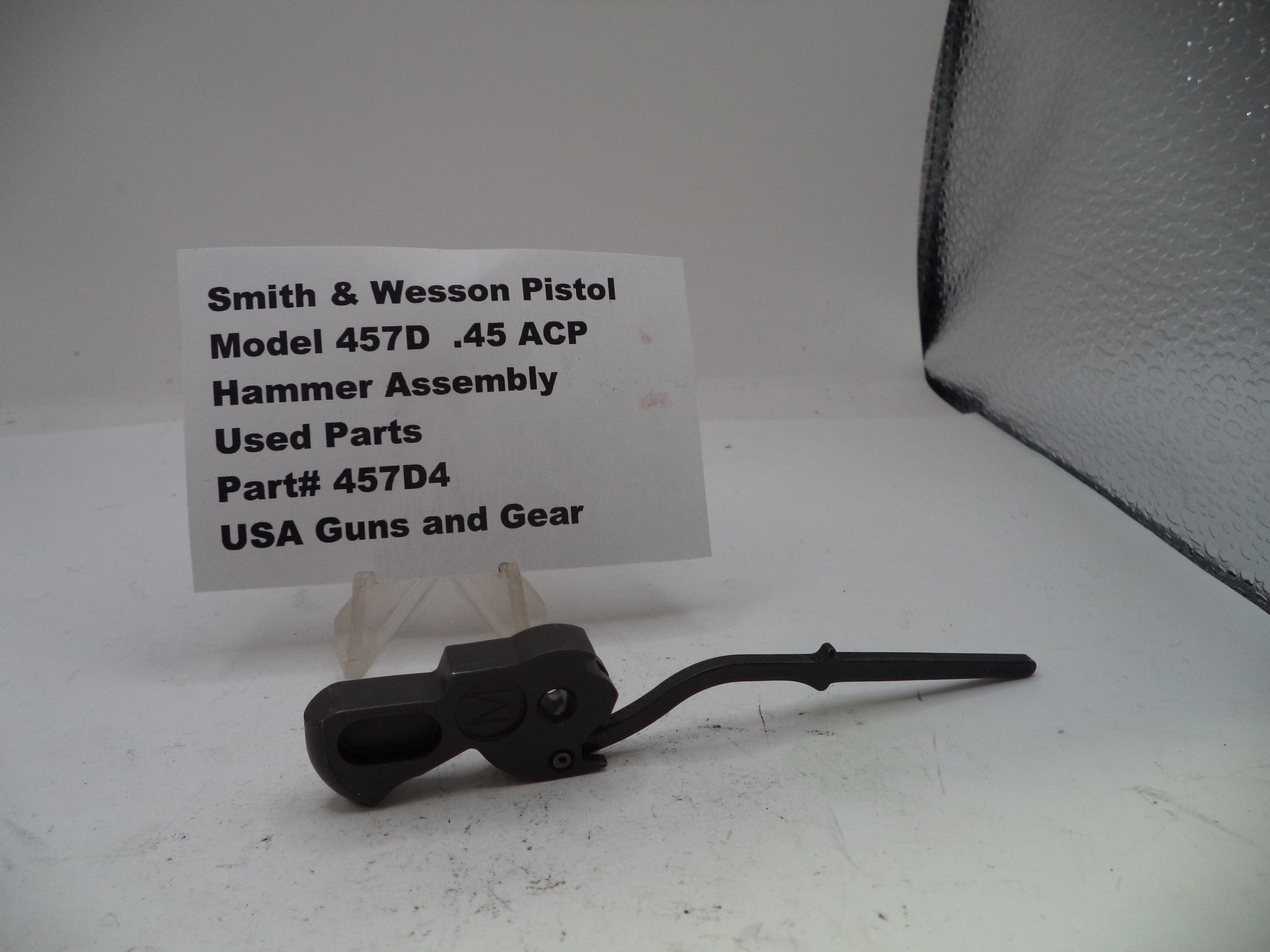 Smith & Wesson pistol hammer assembly on a white surface with a label.