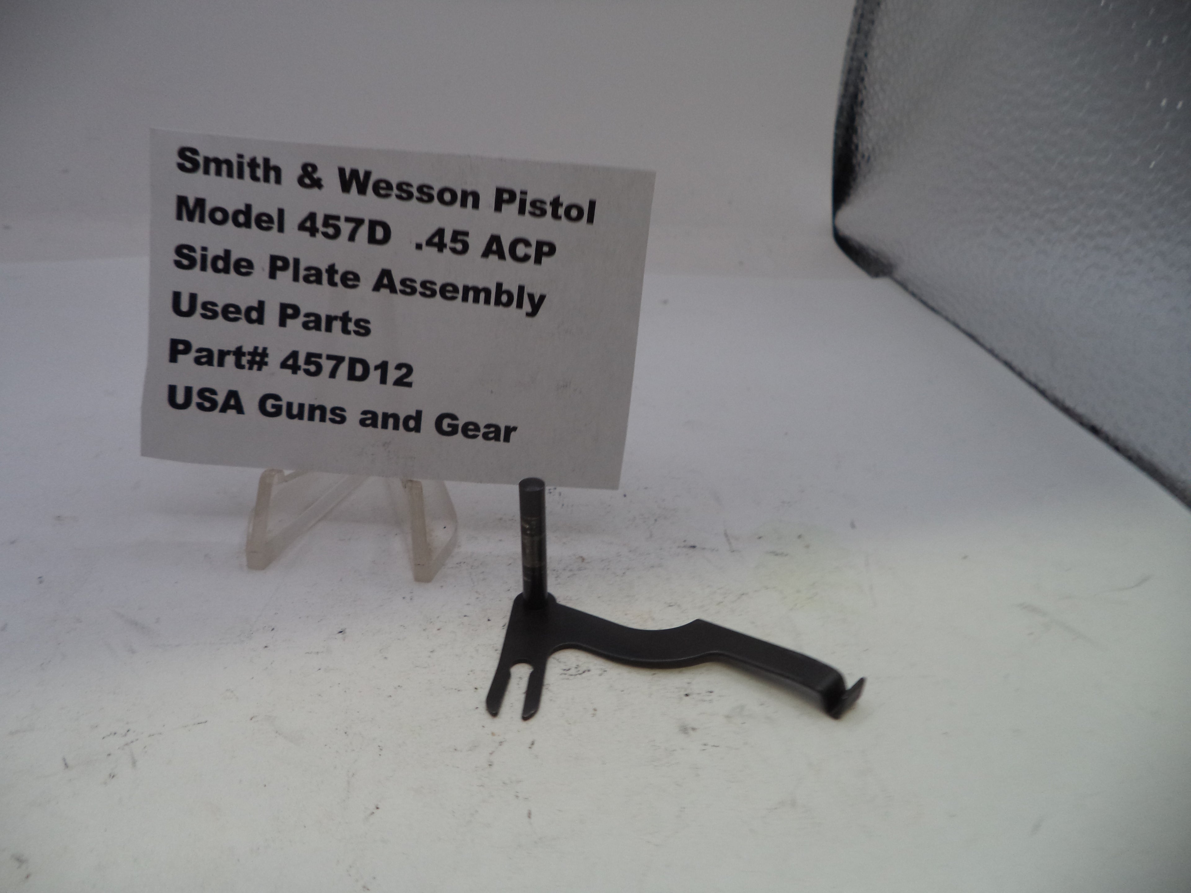 Black Smith & Wesson pistol side plate assembly on a white background with a label.