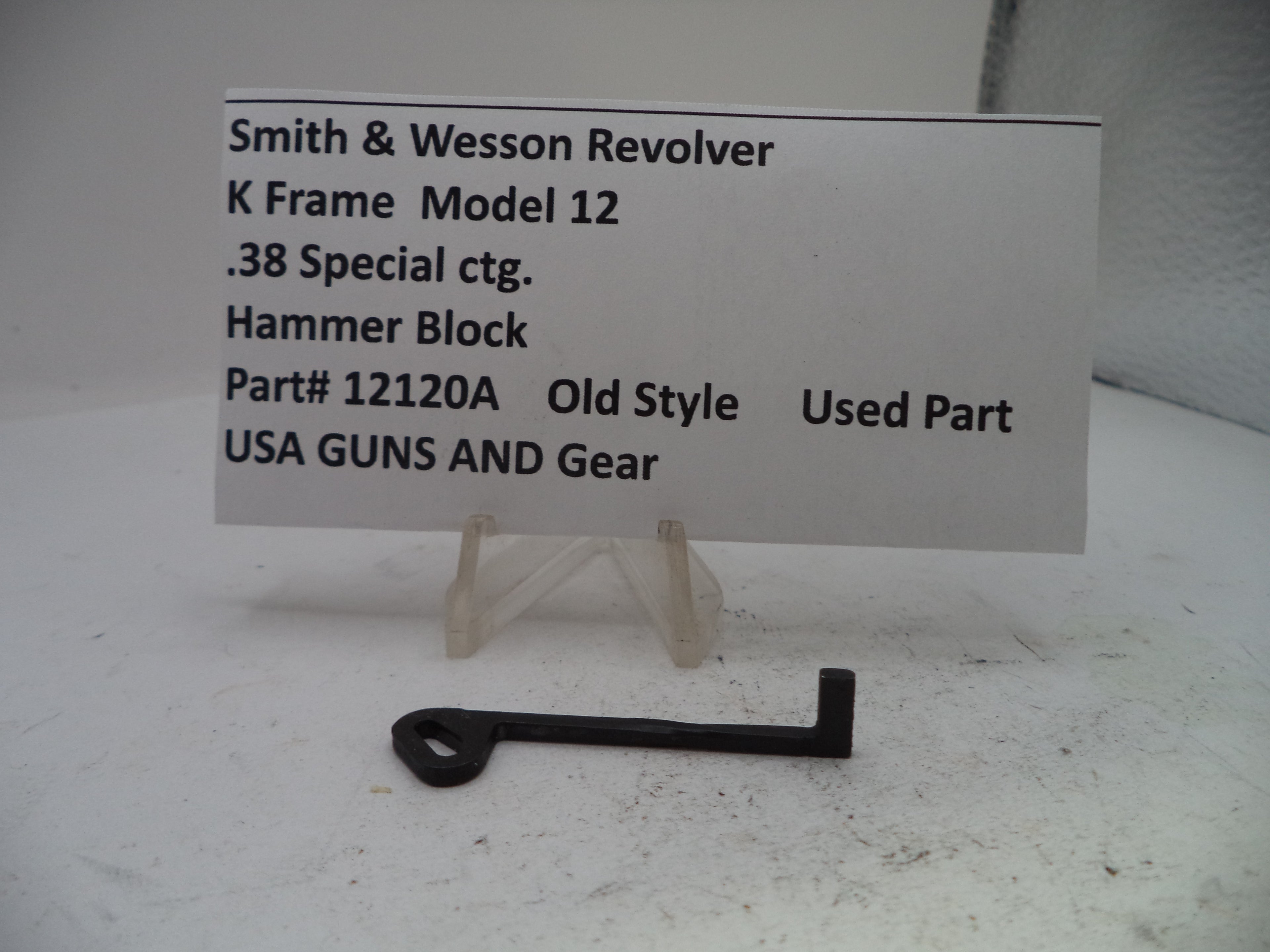 A small black metal hammer block part displayed on a clear stand in front of a white label card that reads 'Smith & Wesson Revolver K Frame Model 12 .38 Special ctg.