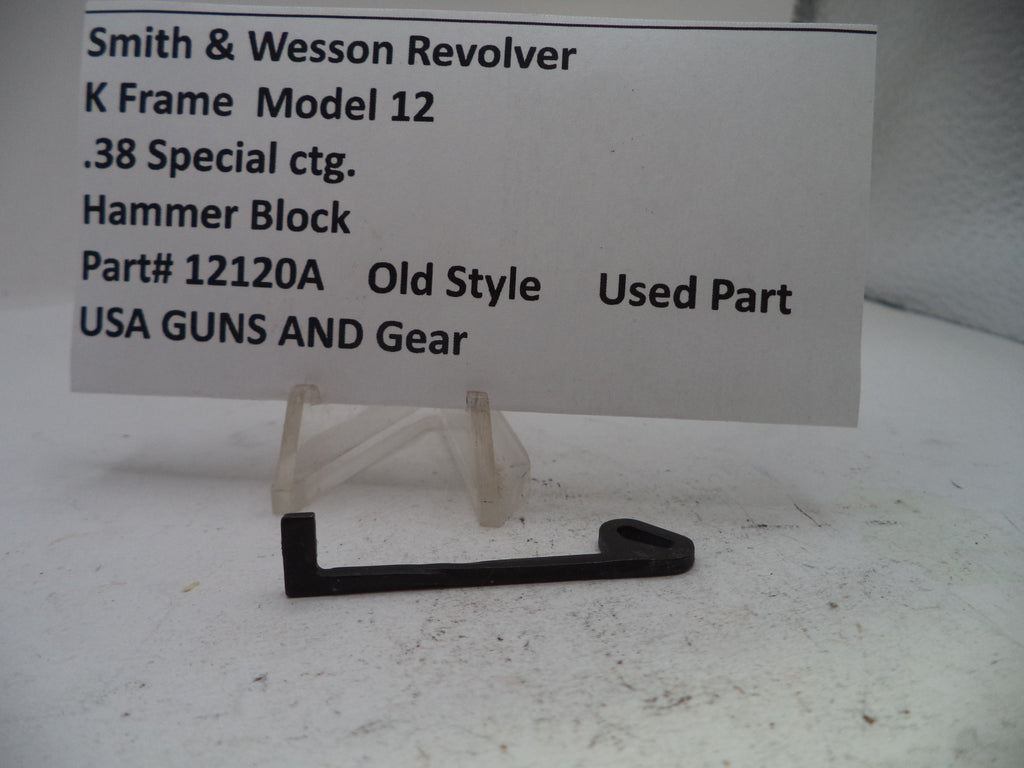 A small black metal hammer block part displayed on a clear plastic stand in front of a white sign that reads 'Smith & Wesson Revolver K Frame Model 12 .38 Special ctg.