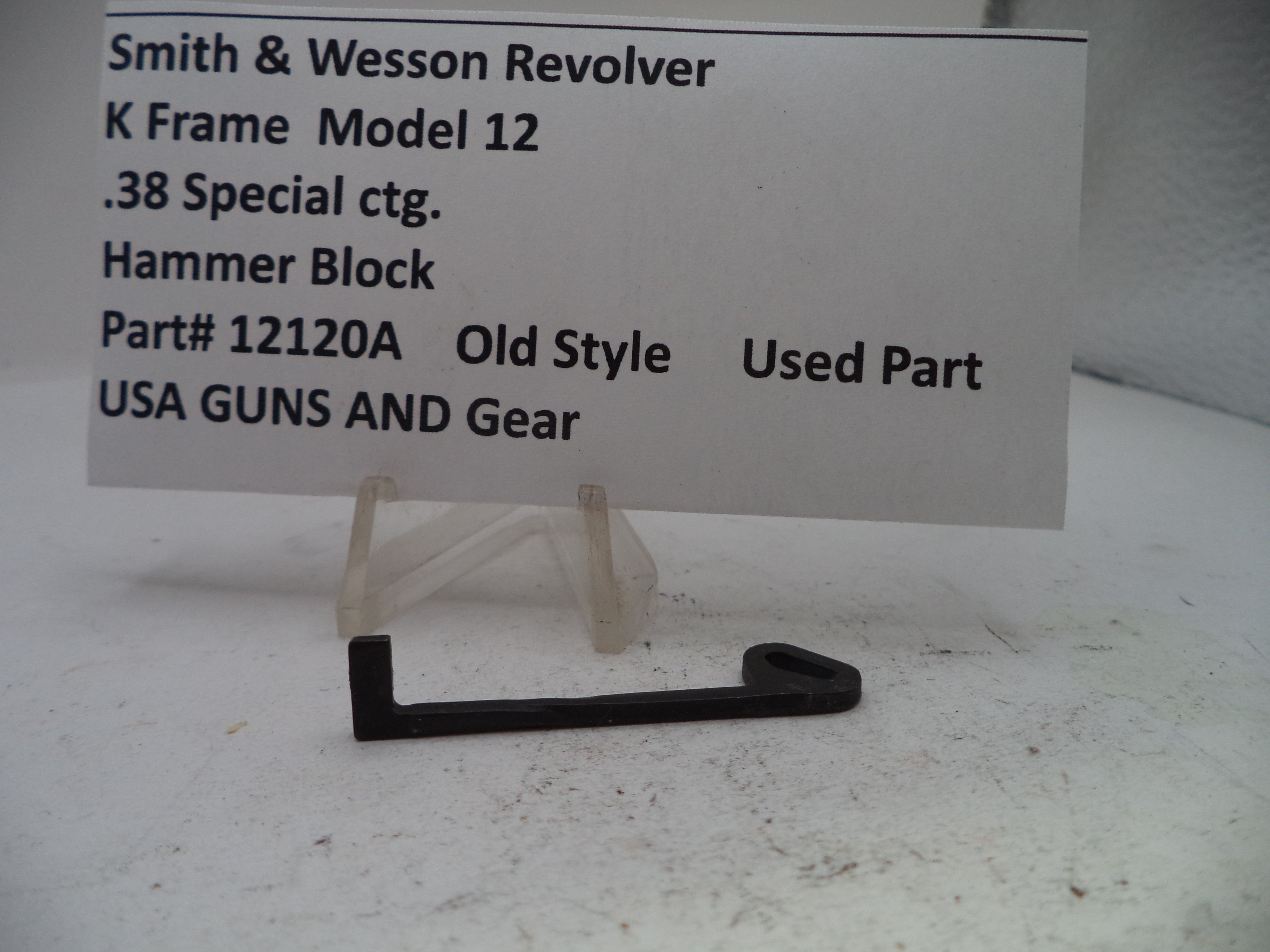 A small black metal hammer block part displayed on a clear plastic stand in front of a white sign that reads 'Smith & Wesson Revolver K Frame Model 12 .38 Special ctg.