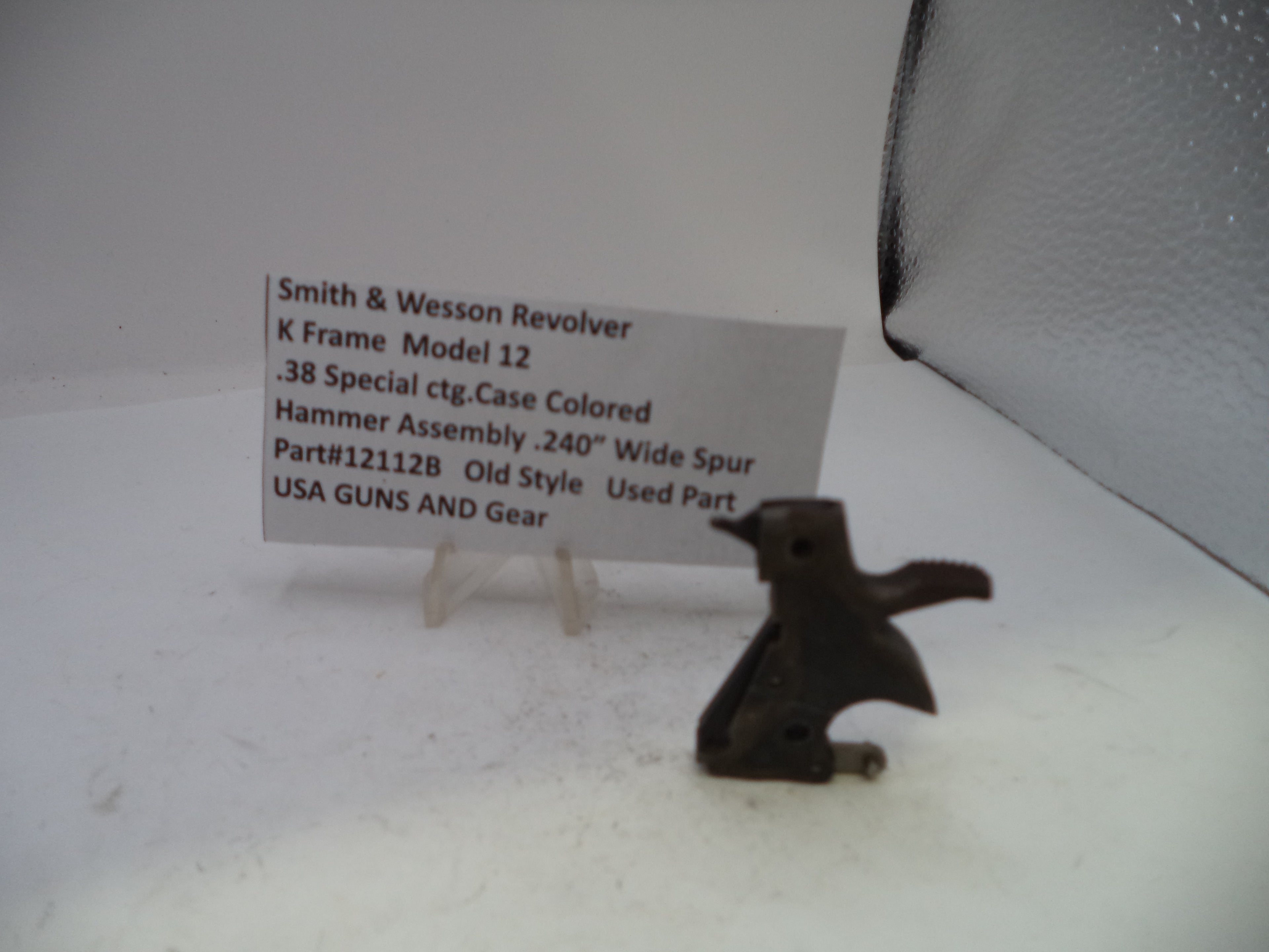 A small dark metal hammer assembly part displayed on a clear stand next to a printed label card for a Smith & Wesson Revolver hammer assembly, photographed against a plain light background.