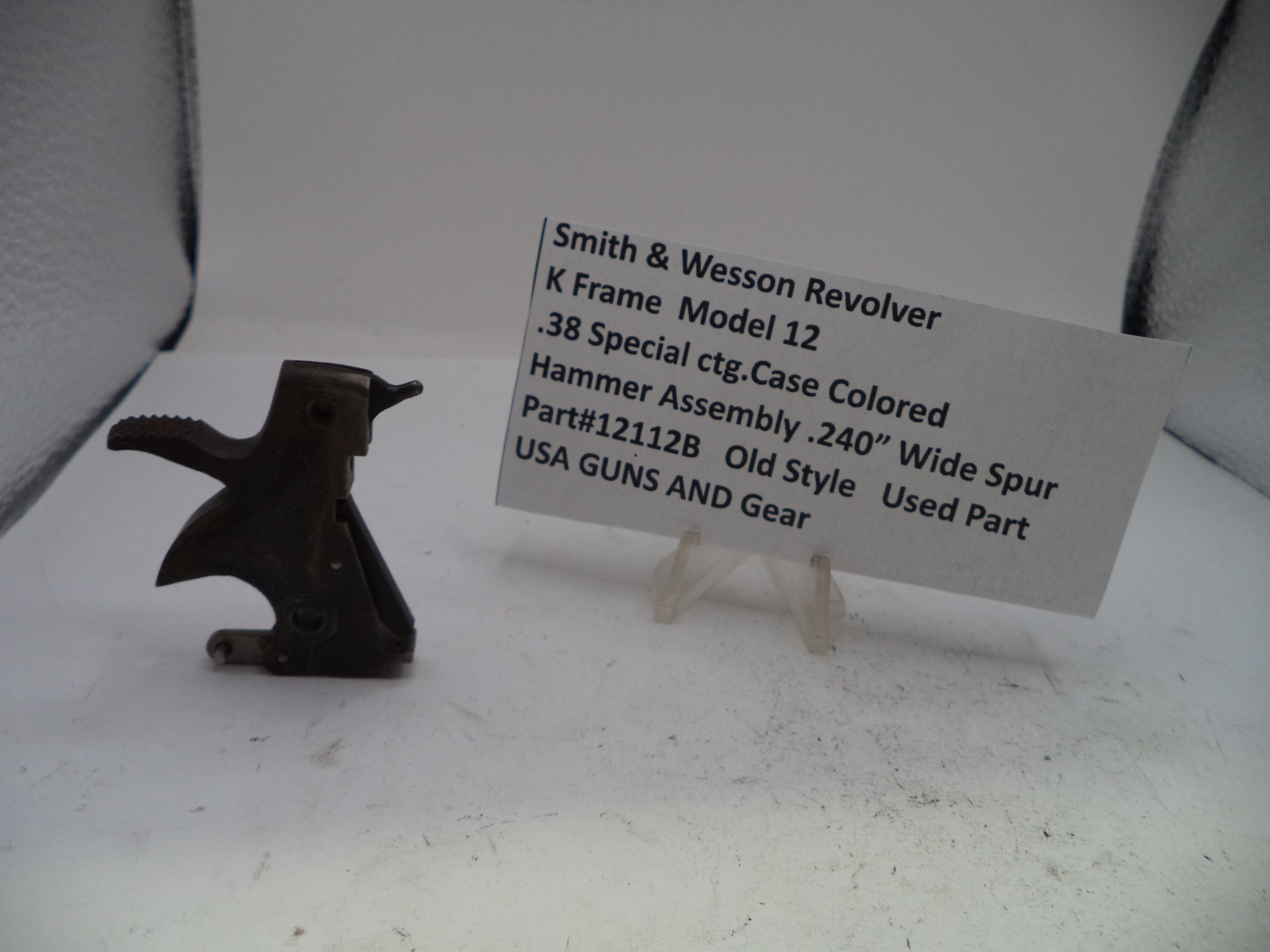 A used metal hammer assembly for a Smith & Wesson revolver, displayed next to a white paper label card on a white surface.