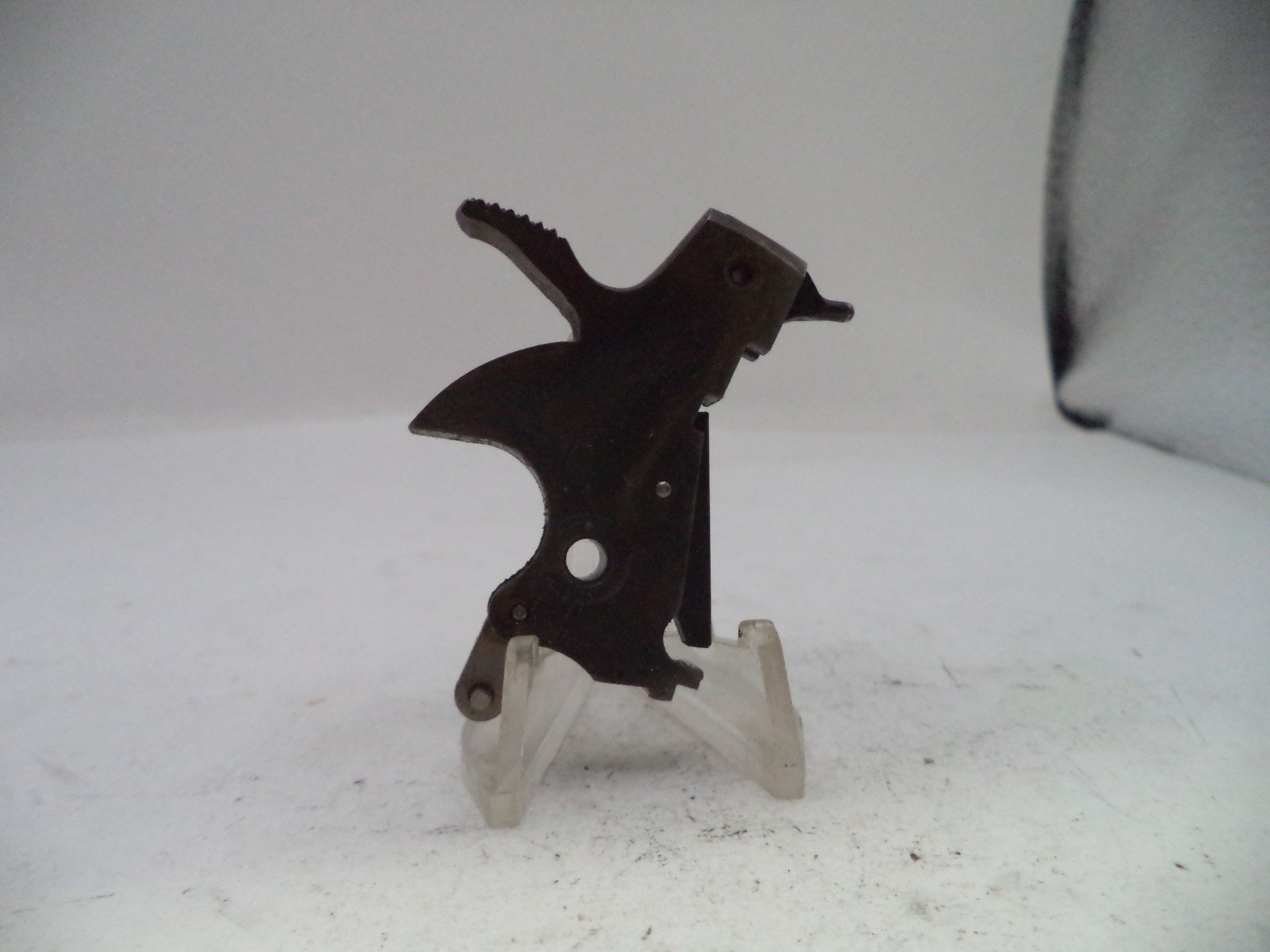 A small dark metal mechanical part or tool component displayed upright on a clear plastic stand against a plain light background.