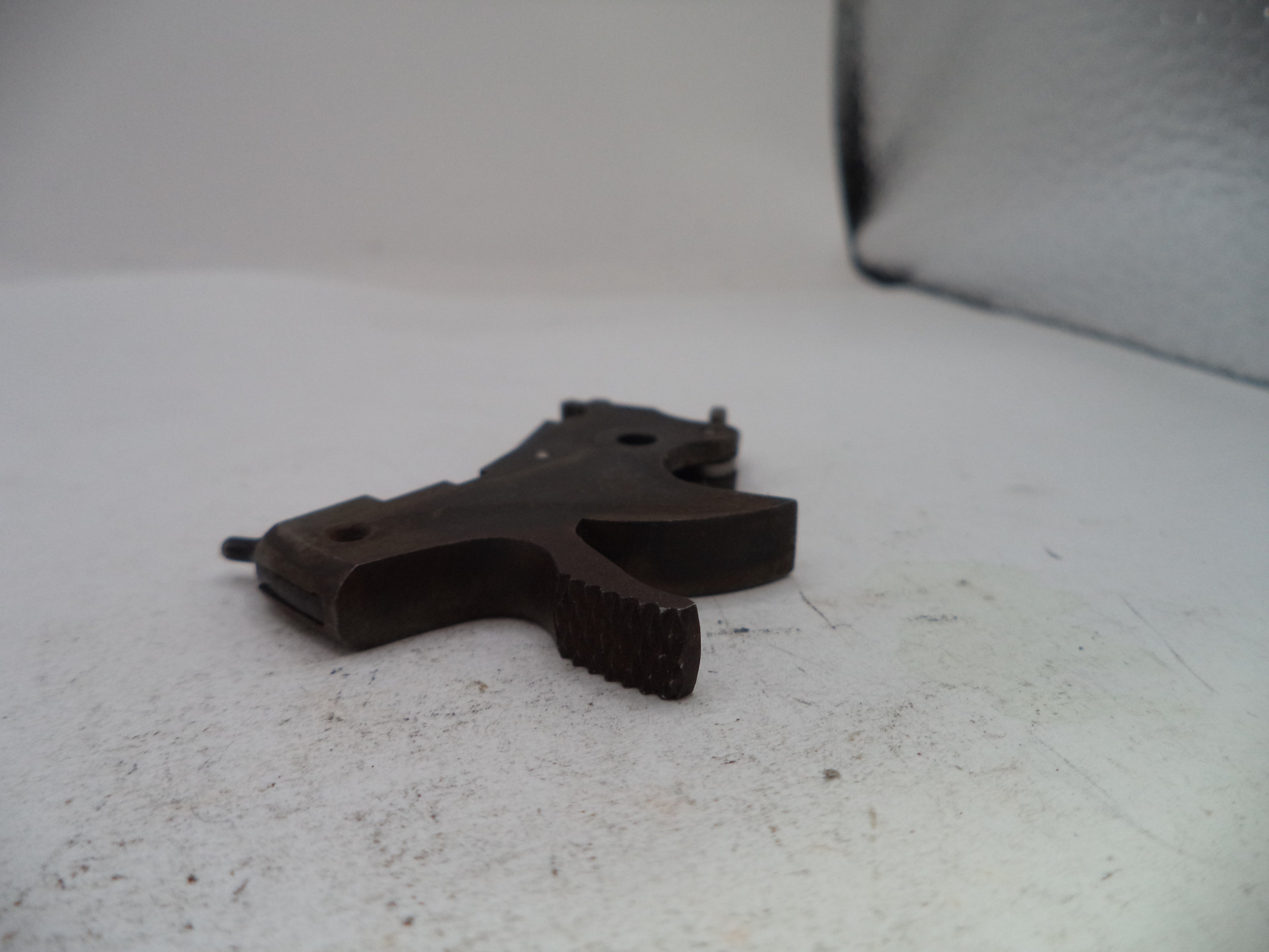 A dark metal firearm trigger assembly, likely a receiver trigger or pistol trigger component, resting on a white surface.