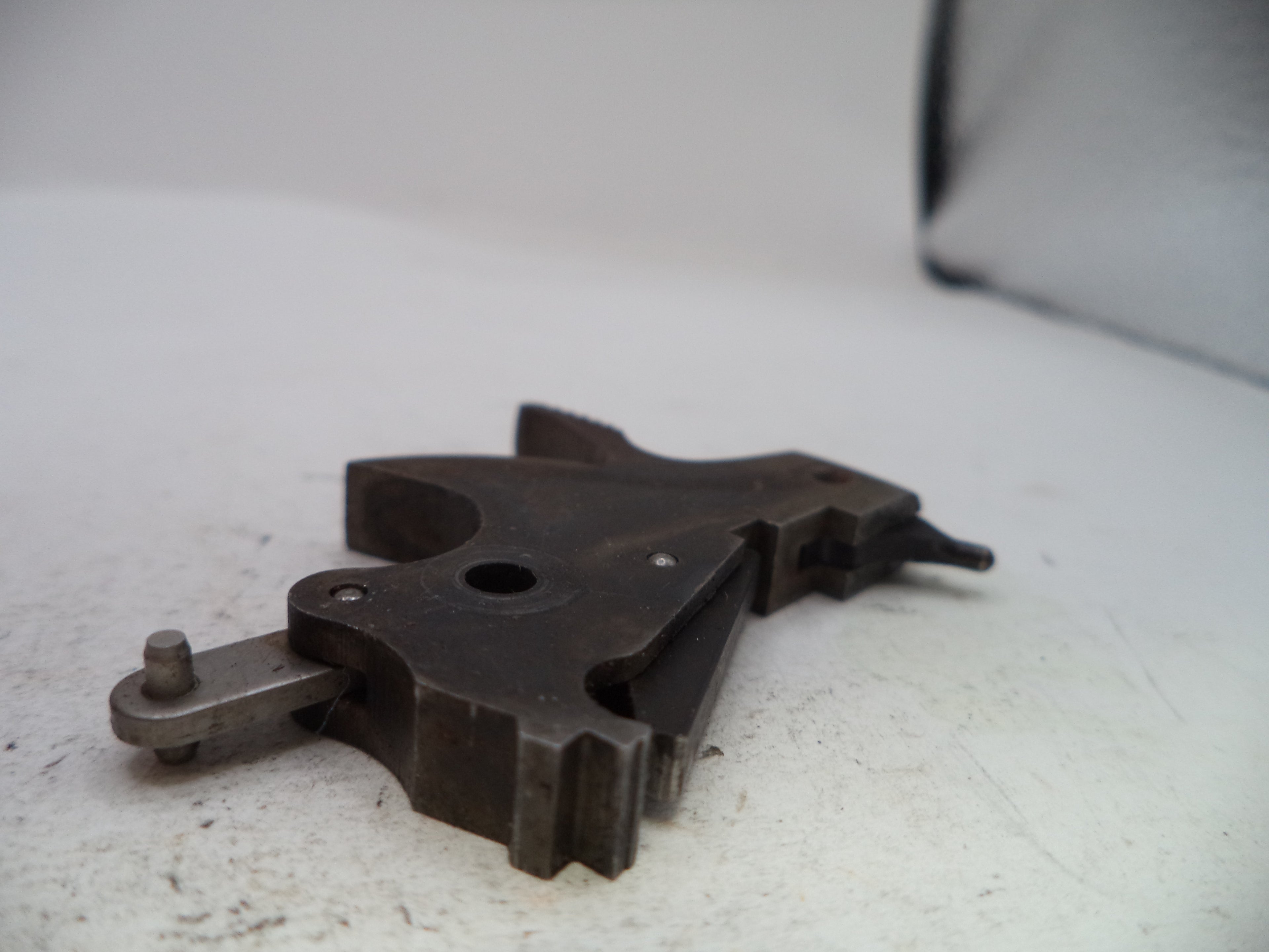 A dark metal mechanical component or tool part with a worn, used surface, photographed on a white surface with a soft-focus background.