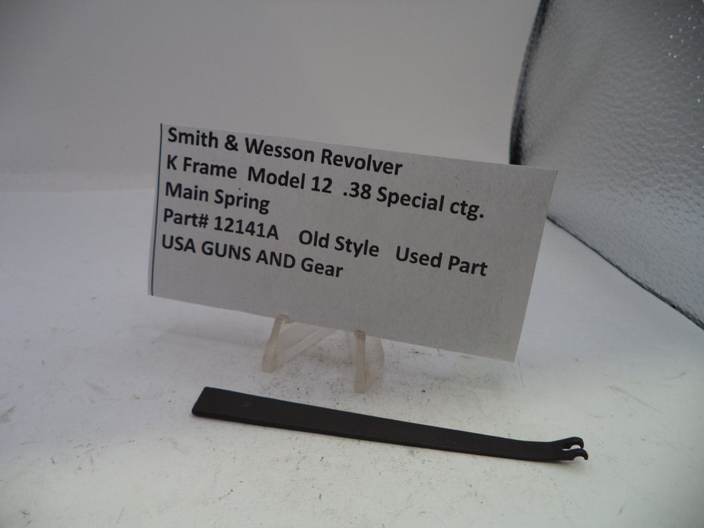 A small black metal spring part displayed on a white surface next to a printed identification card for a Smith & Wesson revolver main spring.