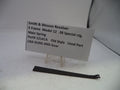 A small black metal spring part displayed on a white surface next to a printed identification card for a Smith & Wesson revolver main spring.
