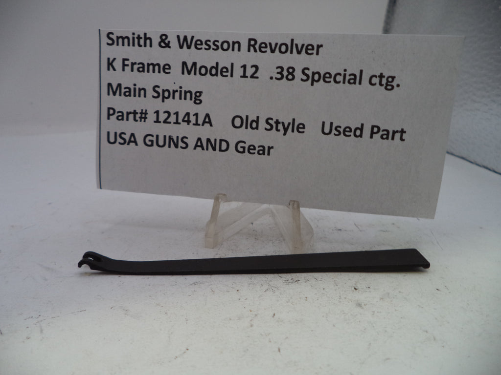 A black metal main spring part for a Smith & Wesson Revolver K Frame Model 12, displayed on a clear plastic stand with an informational paper label showing the part number and manufacturer USA GUNS AND GEAR.