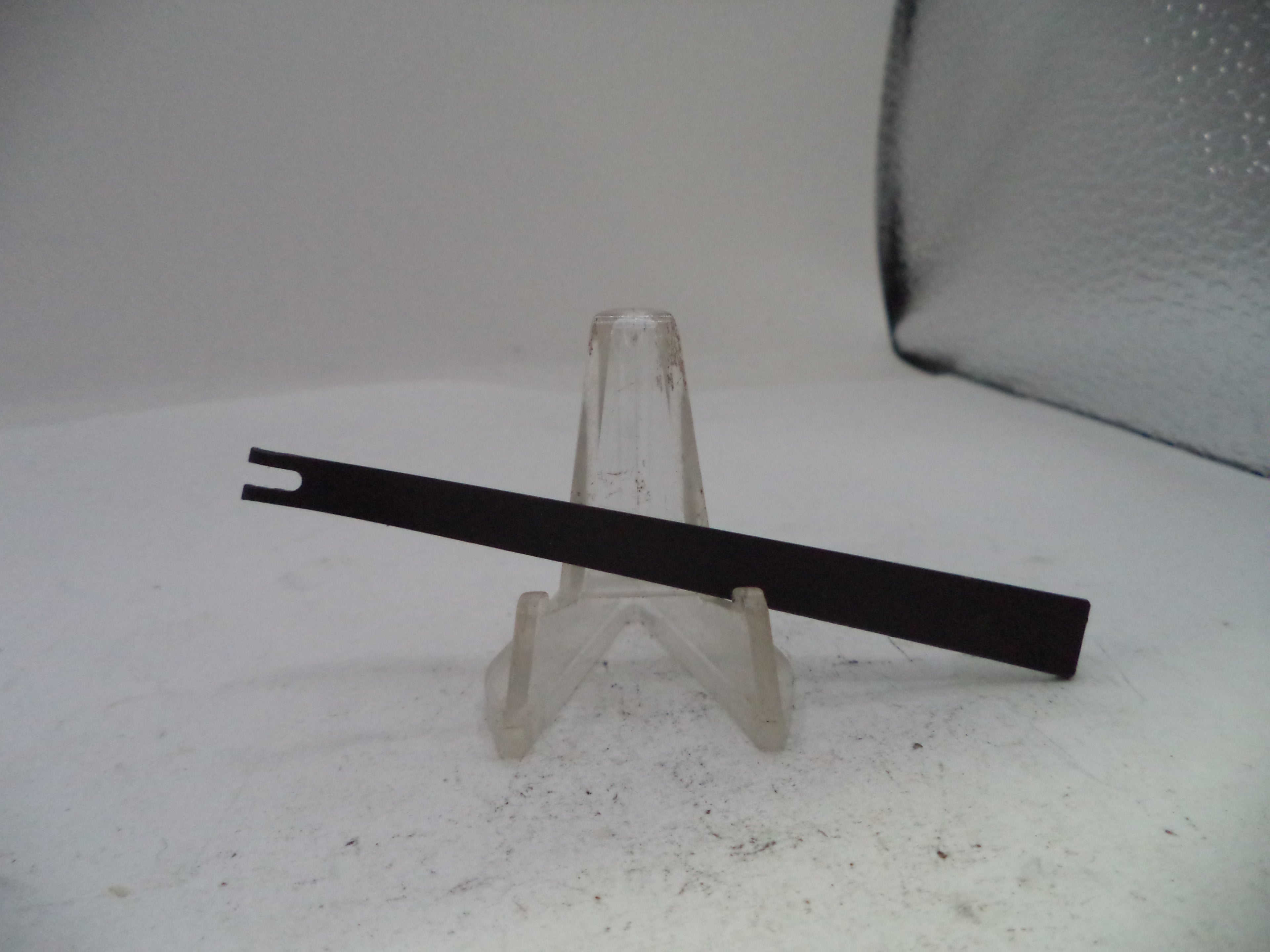 A black metal flathead screwdriver with a double-pronged head, resting on a small clear plastic display stand against a white background.