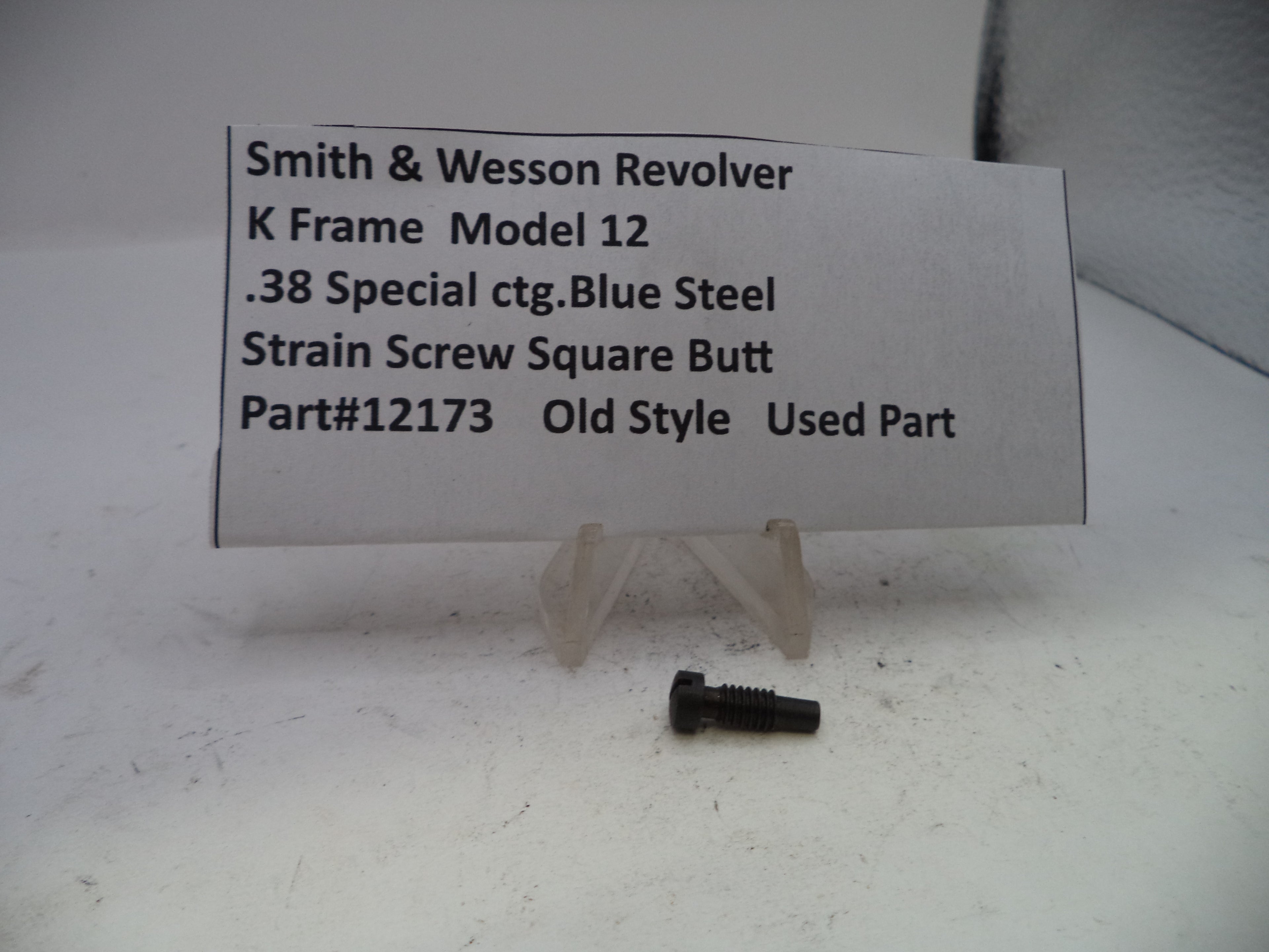 A small used blue steel screw for a Smith & Wesson revolver K frame, displayed on a clear plastic stand next to a white paper label with product details.