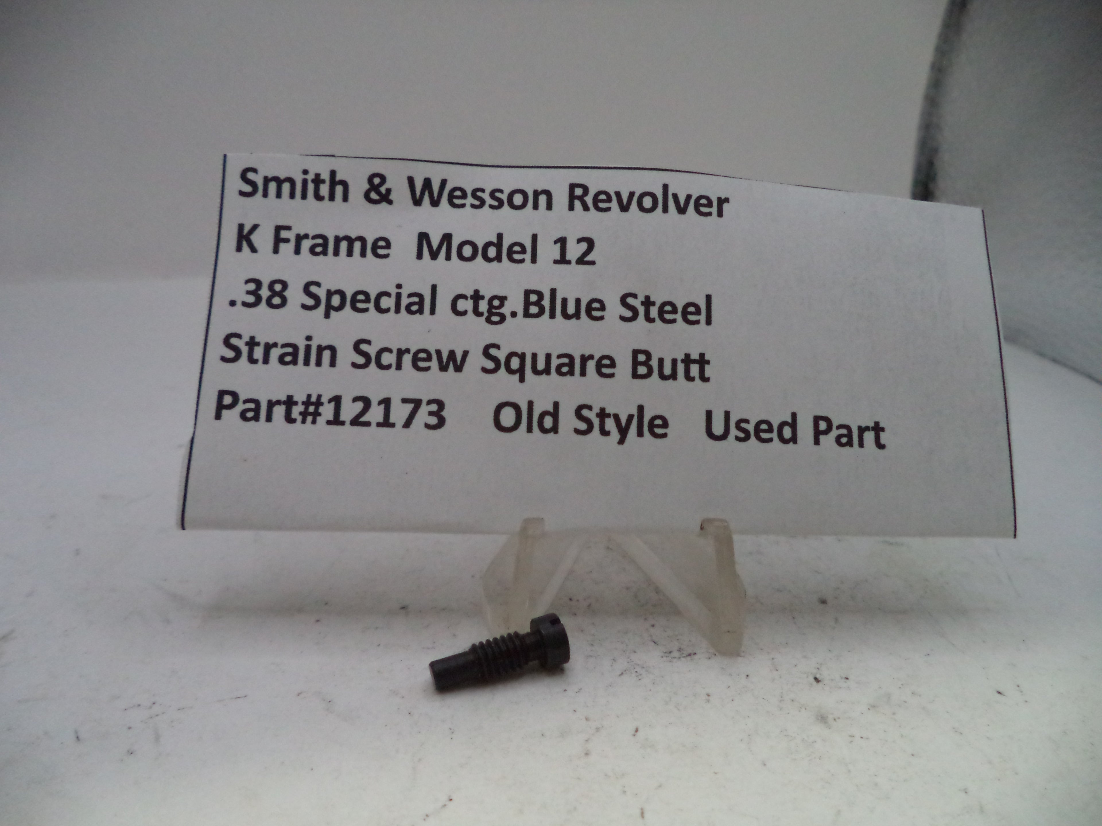 A small black metal strain screw for a Smith & Wesson revolver, displayed on a clear plastic stand next to a white paper label with product details.