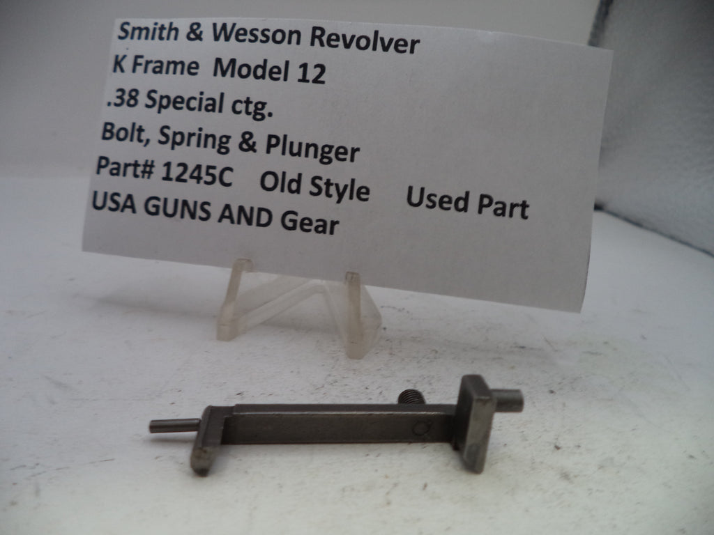 A used metal bolt, spring, and plunger part for a Smith & Wesson revolver, displayed on a small clear stand in front of a white paper label card.