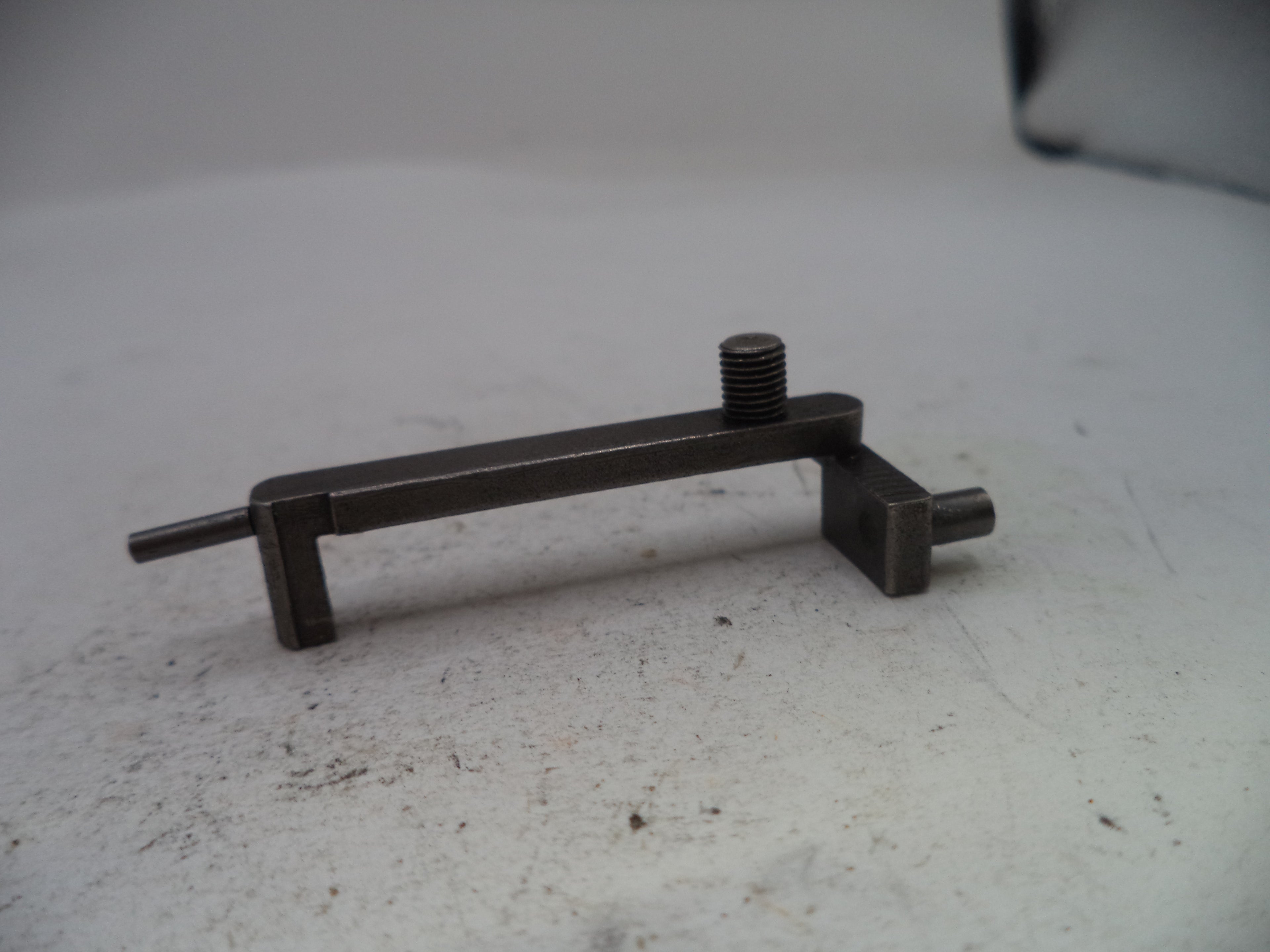 A small dark metal mechanical component with a threaded screw and flat shafts, resting on a light gray surface.