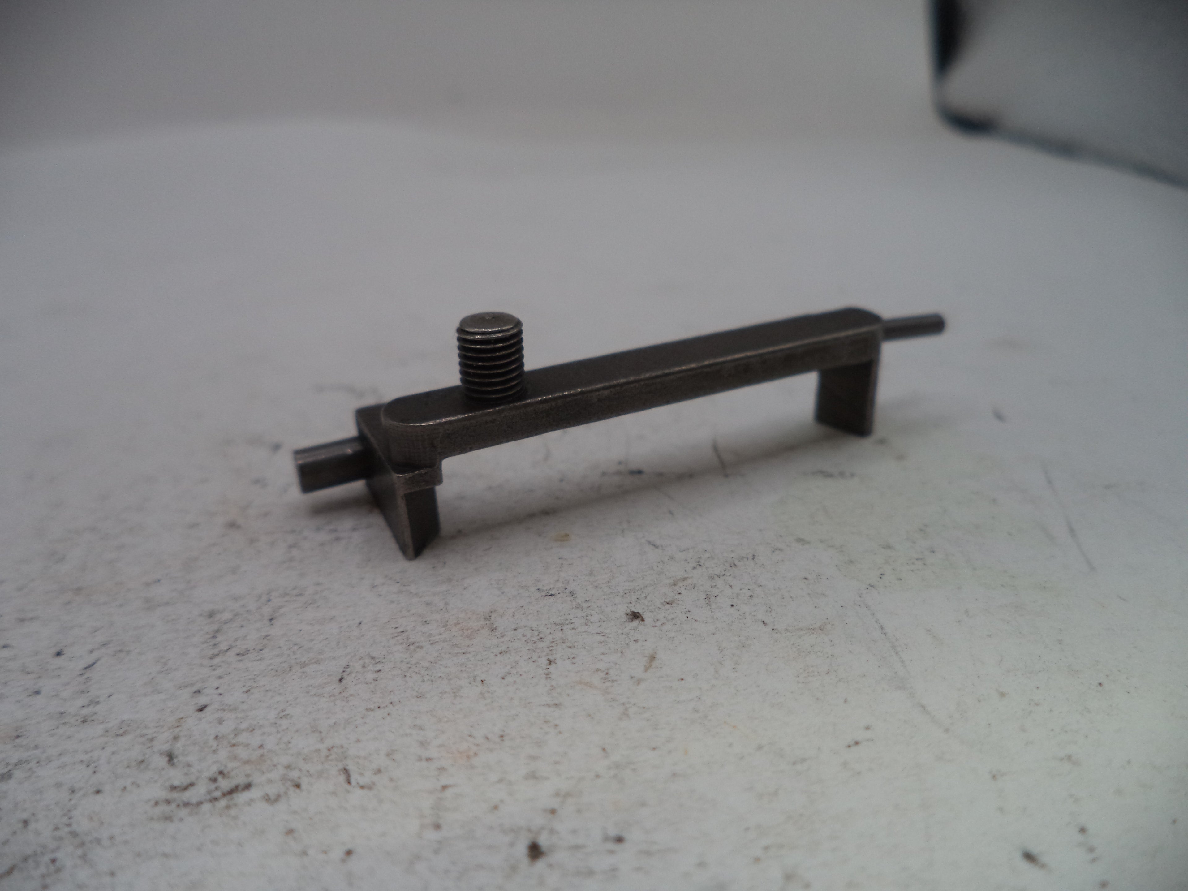 A small dark metal hardware component with a threaded rod and flat bar, photographed on a light gray surface.
