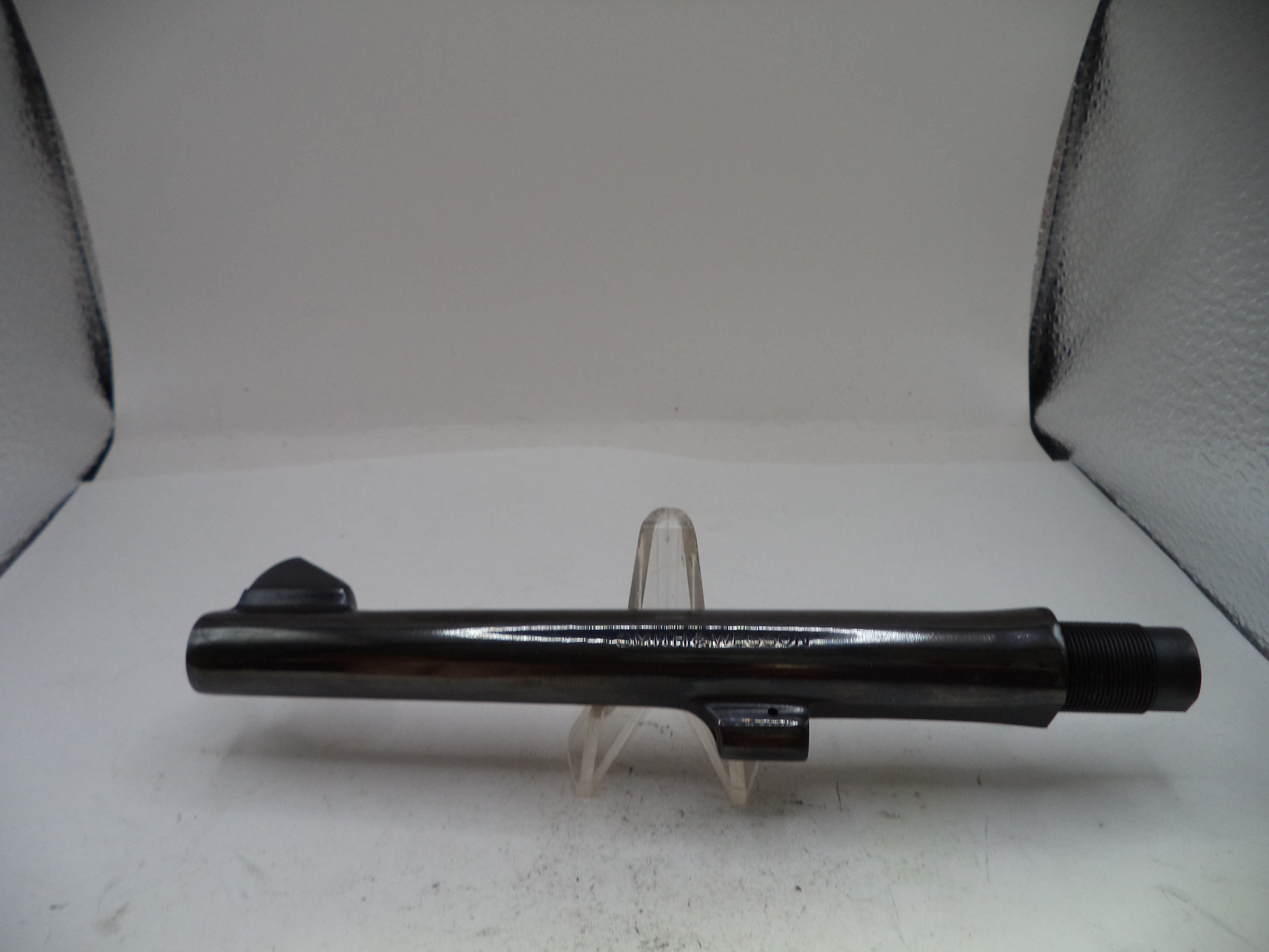 A black metal cylindrical component, likely a firearm barrel or tube, displayed horizontally on a clear plastic stand against a white backdrop with textured black corners.