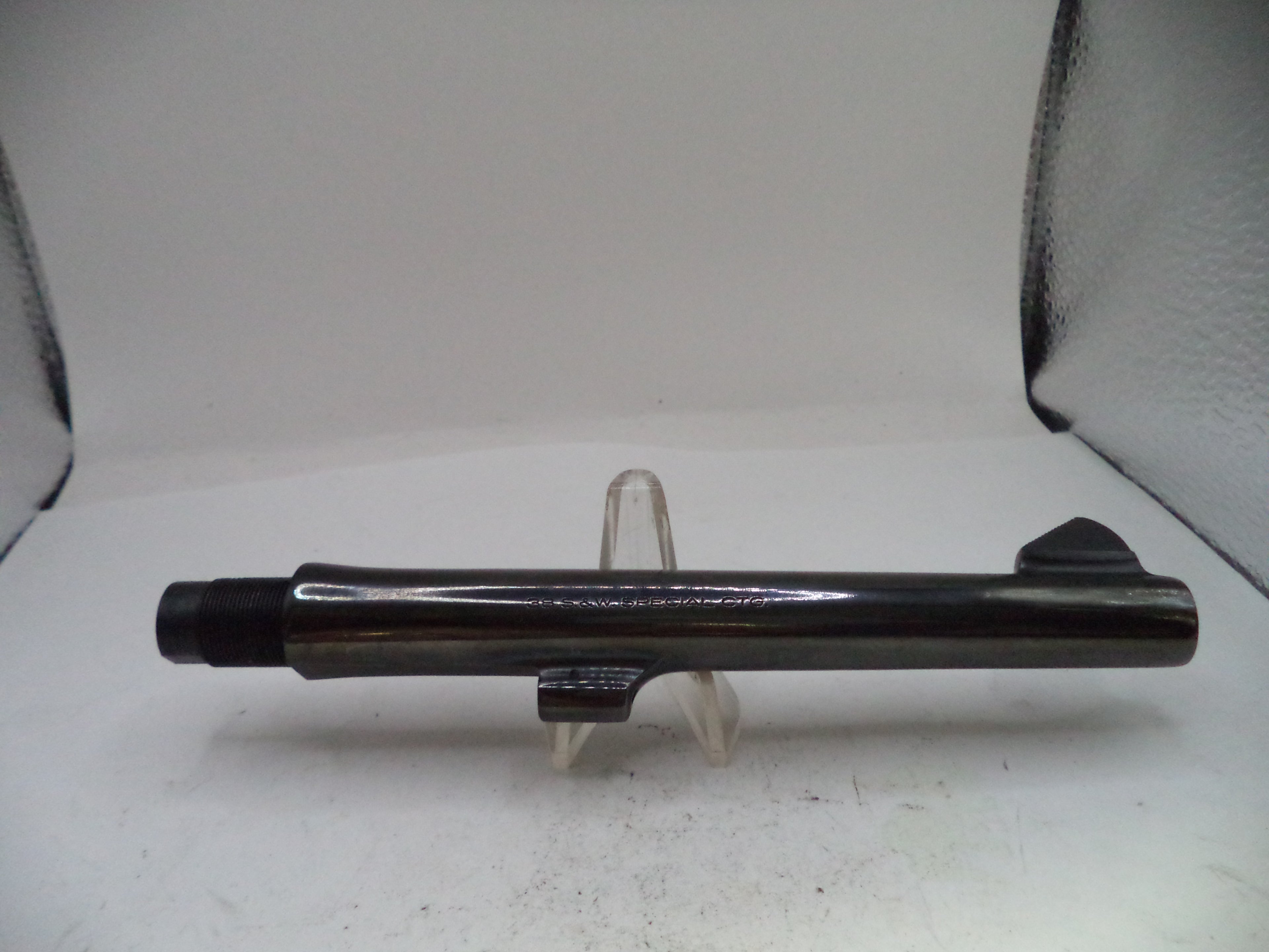 A black metal cylindrical component, likely a firearm barrel or tube, displayed horizontally on a small clear plastic stand against a white backdrop.