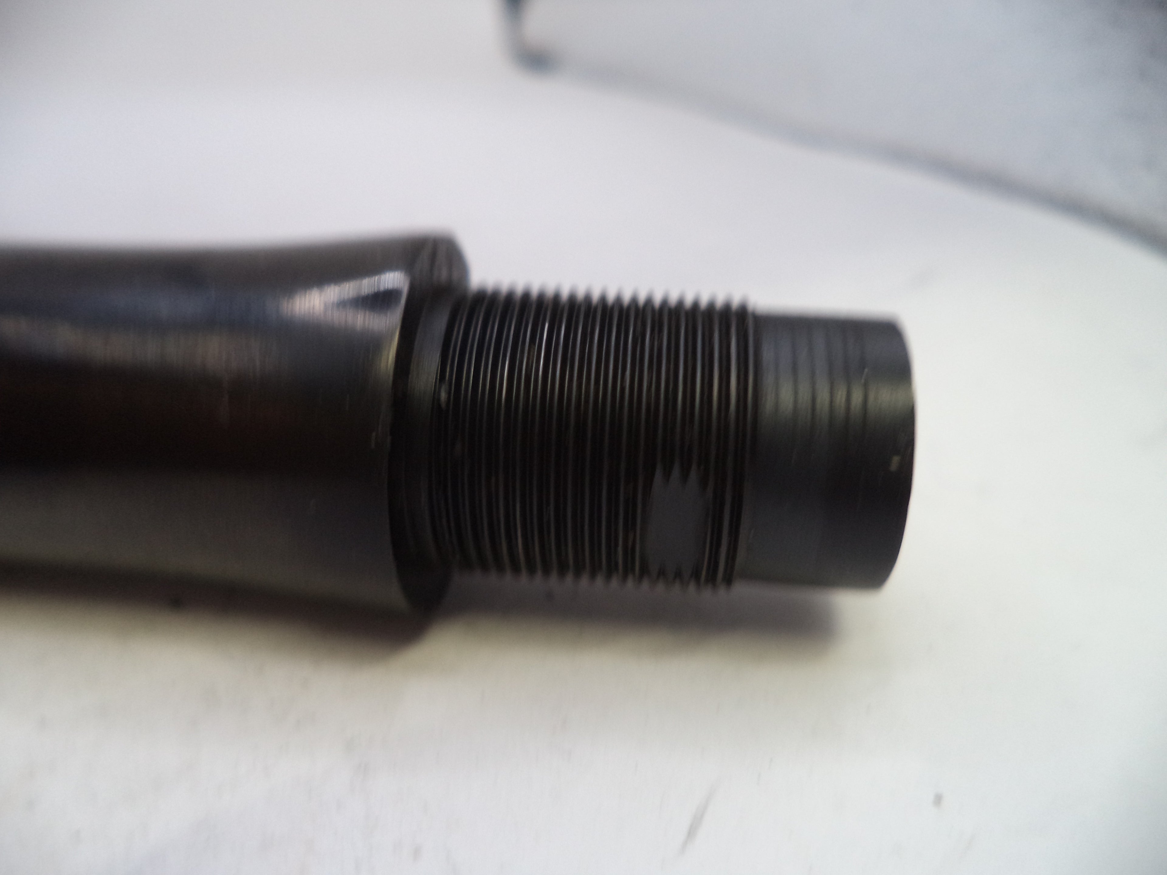 A close-up of a black metal threaded component, possibly a mechanical fitting or valve stem, resting on a white surface.