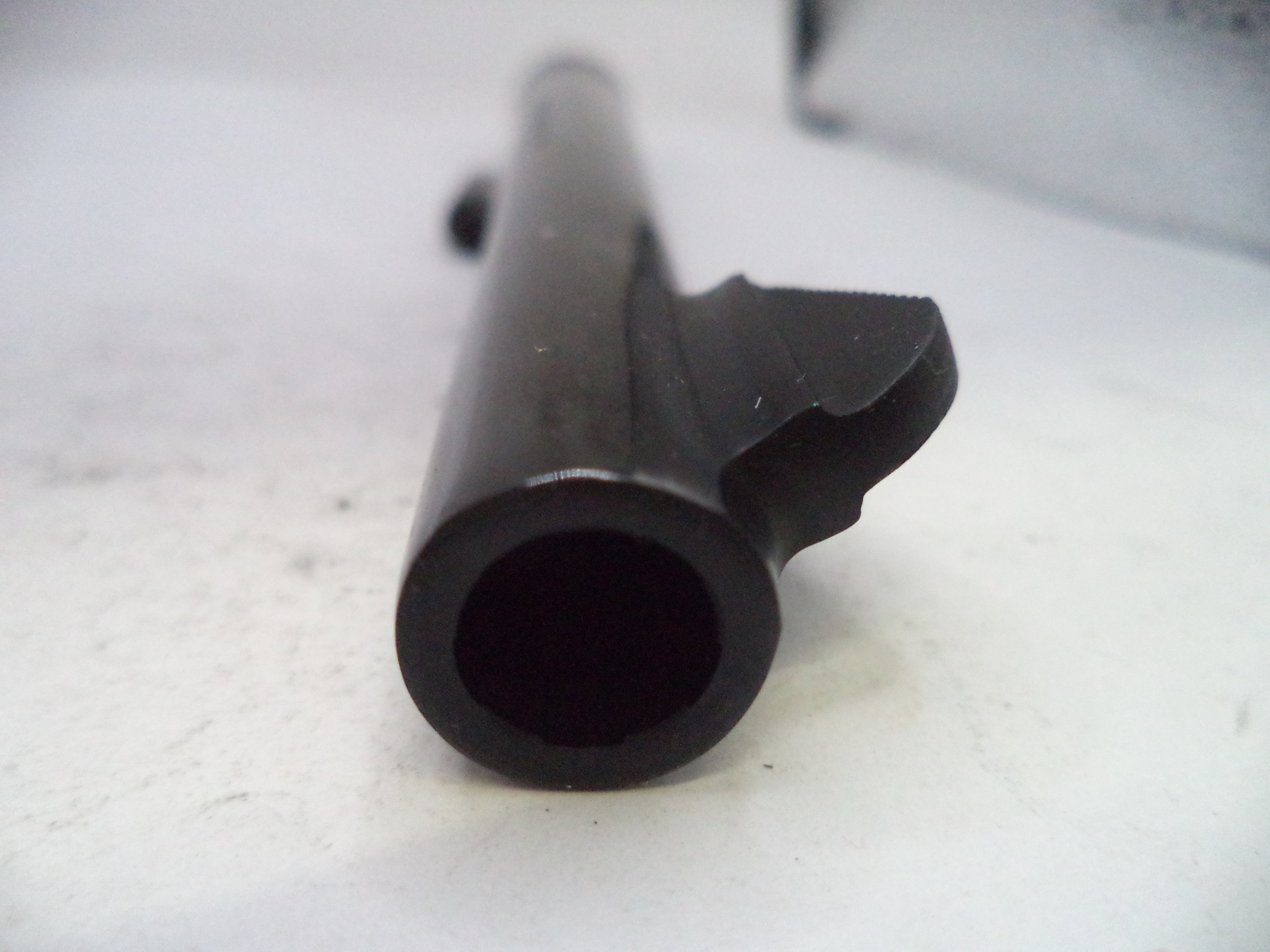 A close-up shot of a black metal cylindrical component, possibly a barrel or tube, with a bracket or clip attached on one side, resting on a light-colored surface.