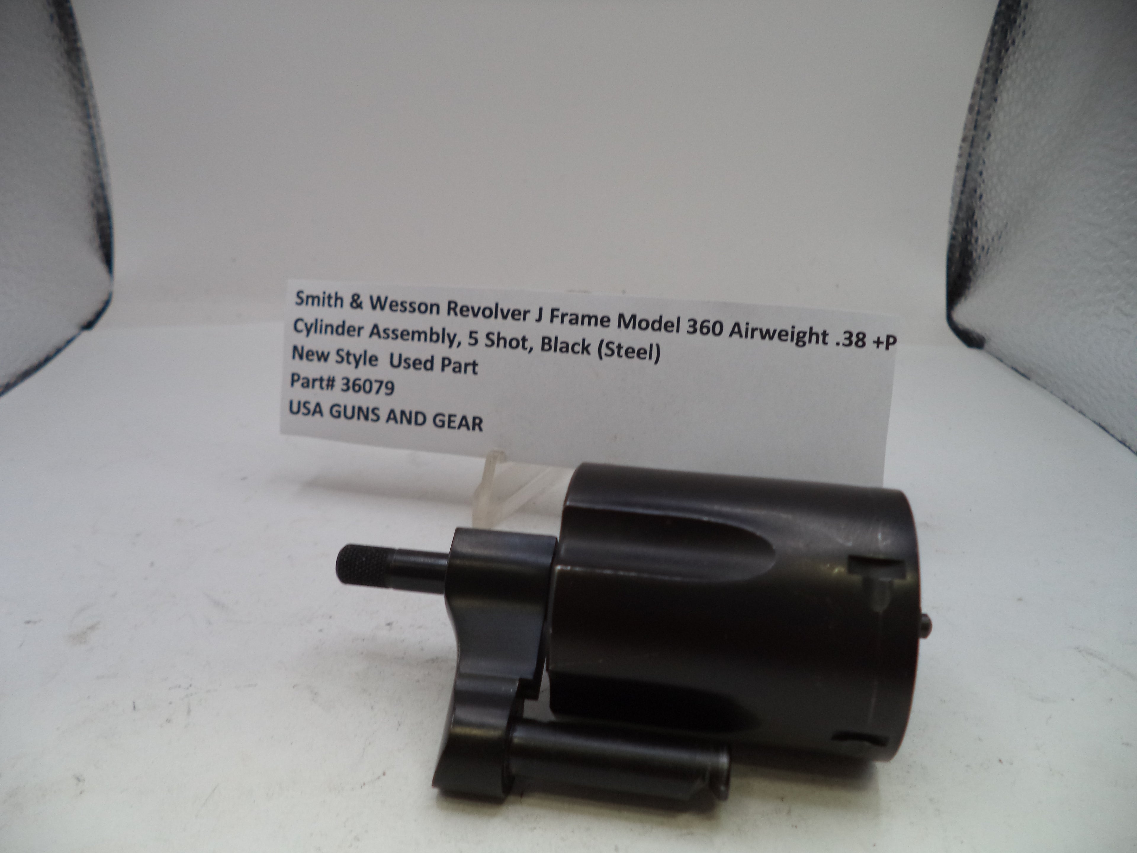 A black Smith & Wesson revolver cylinder assembly part, photographed on a white surface with a handwritten label card behind it.