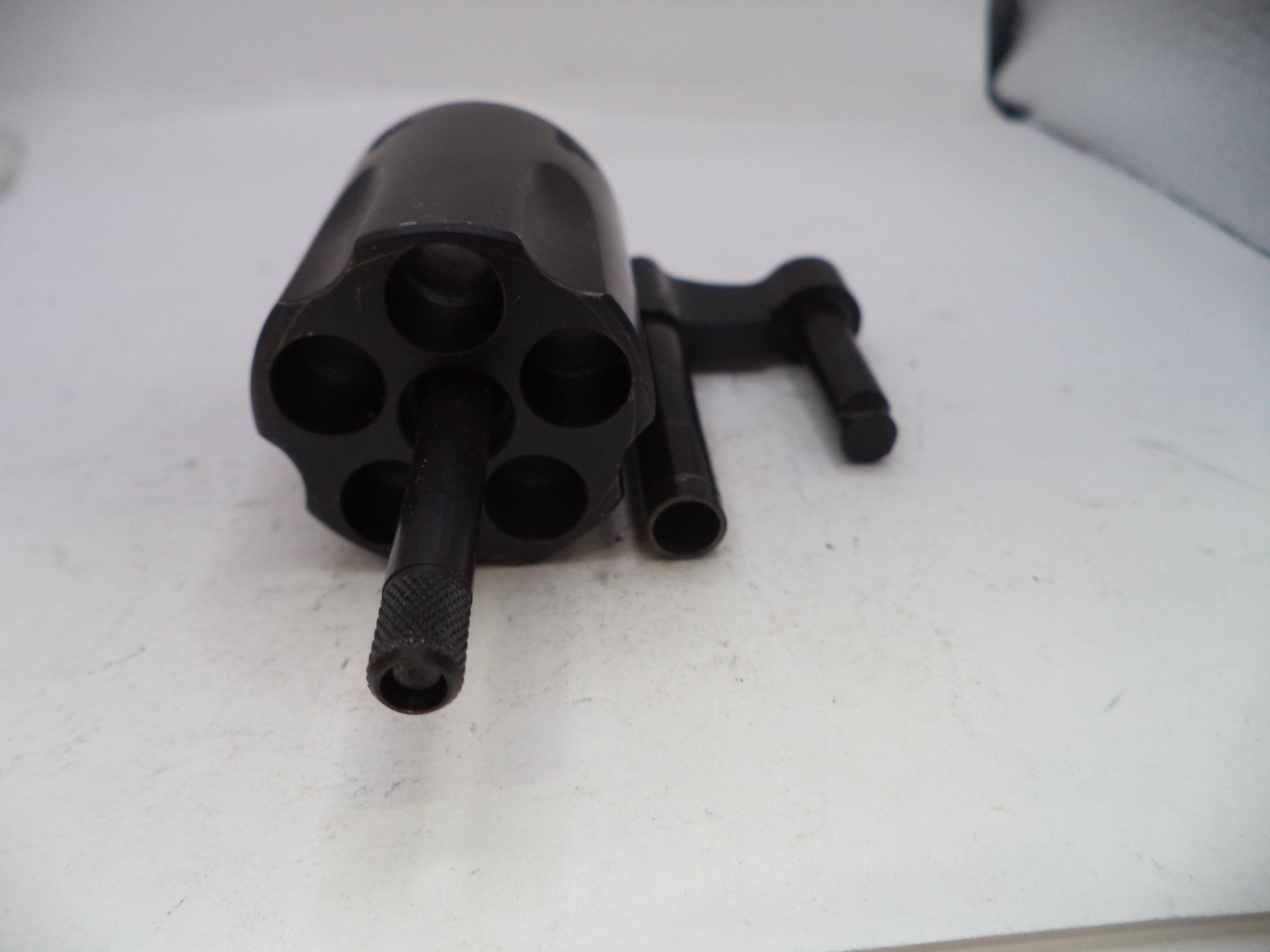 A black metal revolver-style tool or component with a cylindrical opening and a short handle, photographed on a plain light surface.