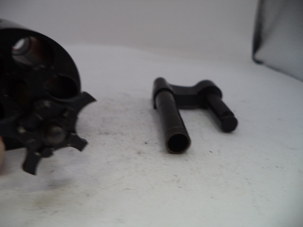 A close-up of a black metal revolver cylinder disassembled to show the internal barrel and a separate black metal pin or bolt component on a white surface.