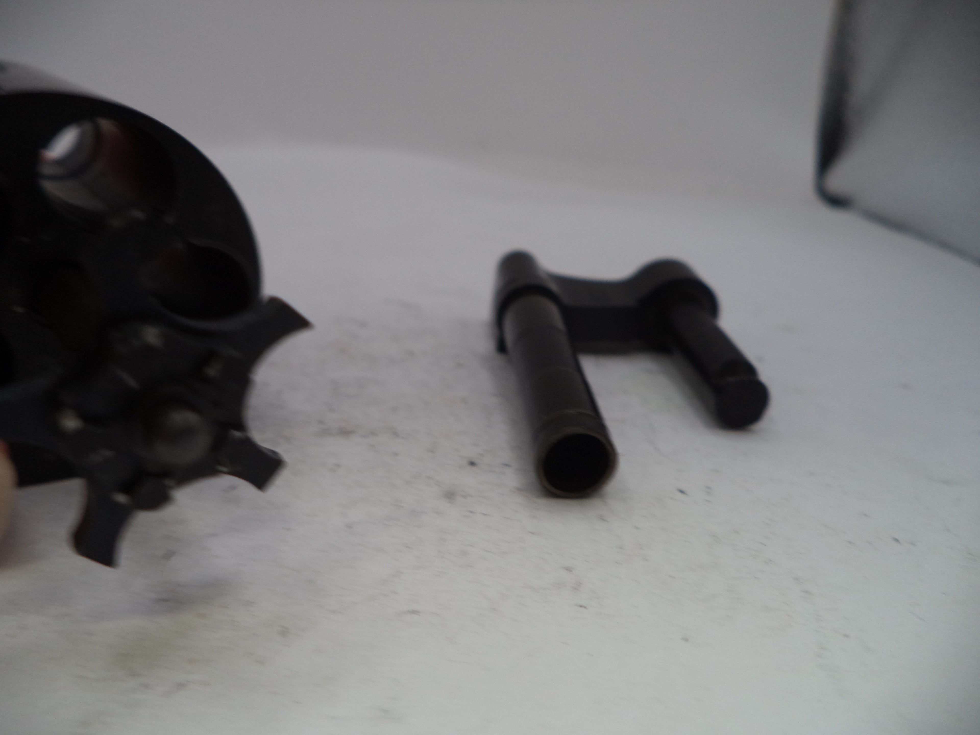 A close-up of a black metal revolver cylinder disassembled to show the internal barrel and a separate black metal pin or bolt component on a white surface.