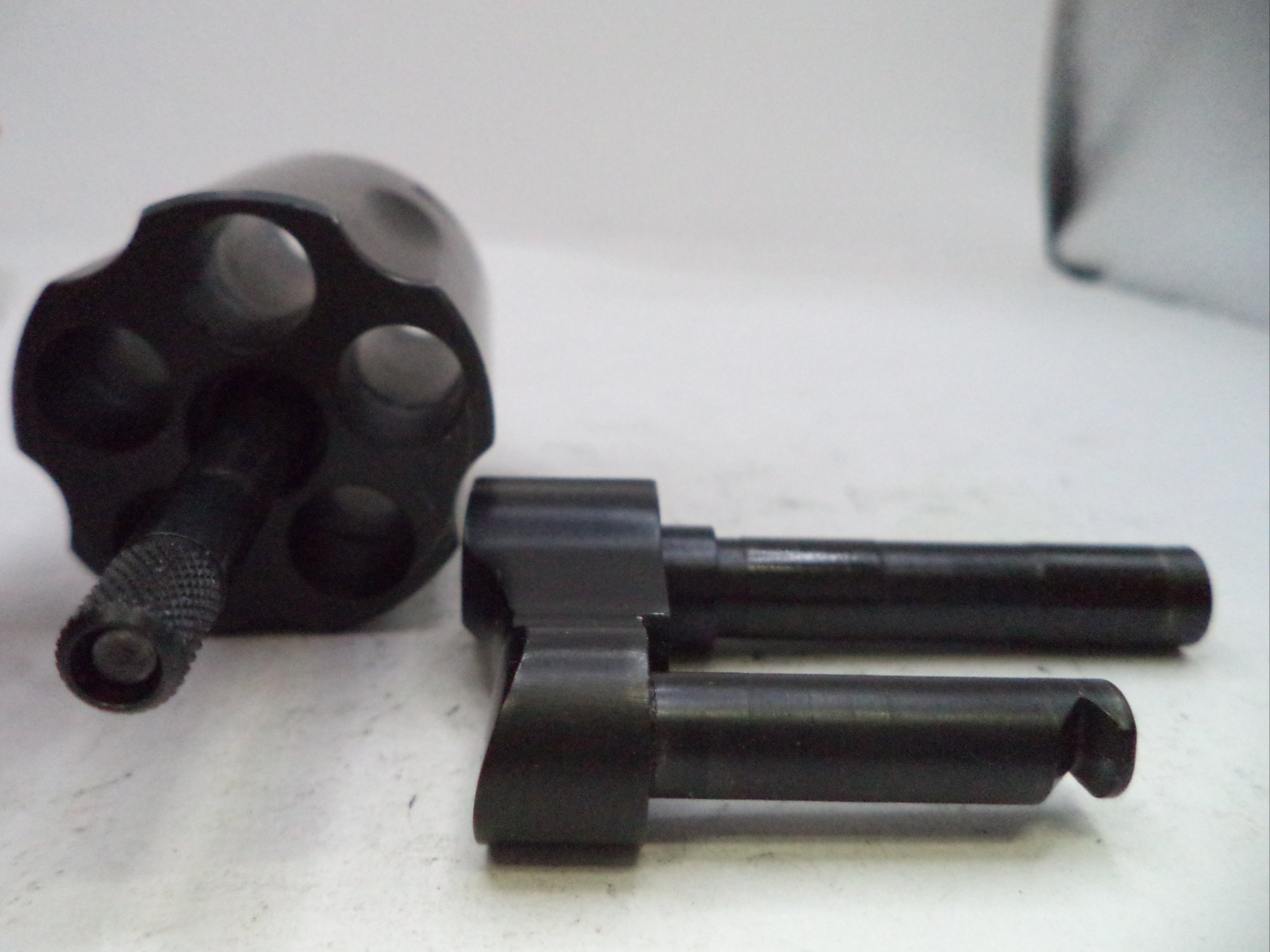 A black metal revolver cylinder disassembled into its components, including a six-cylinder chamber, barrel, and pinion, resting on a white surface.