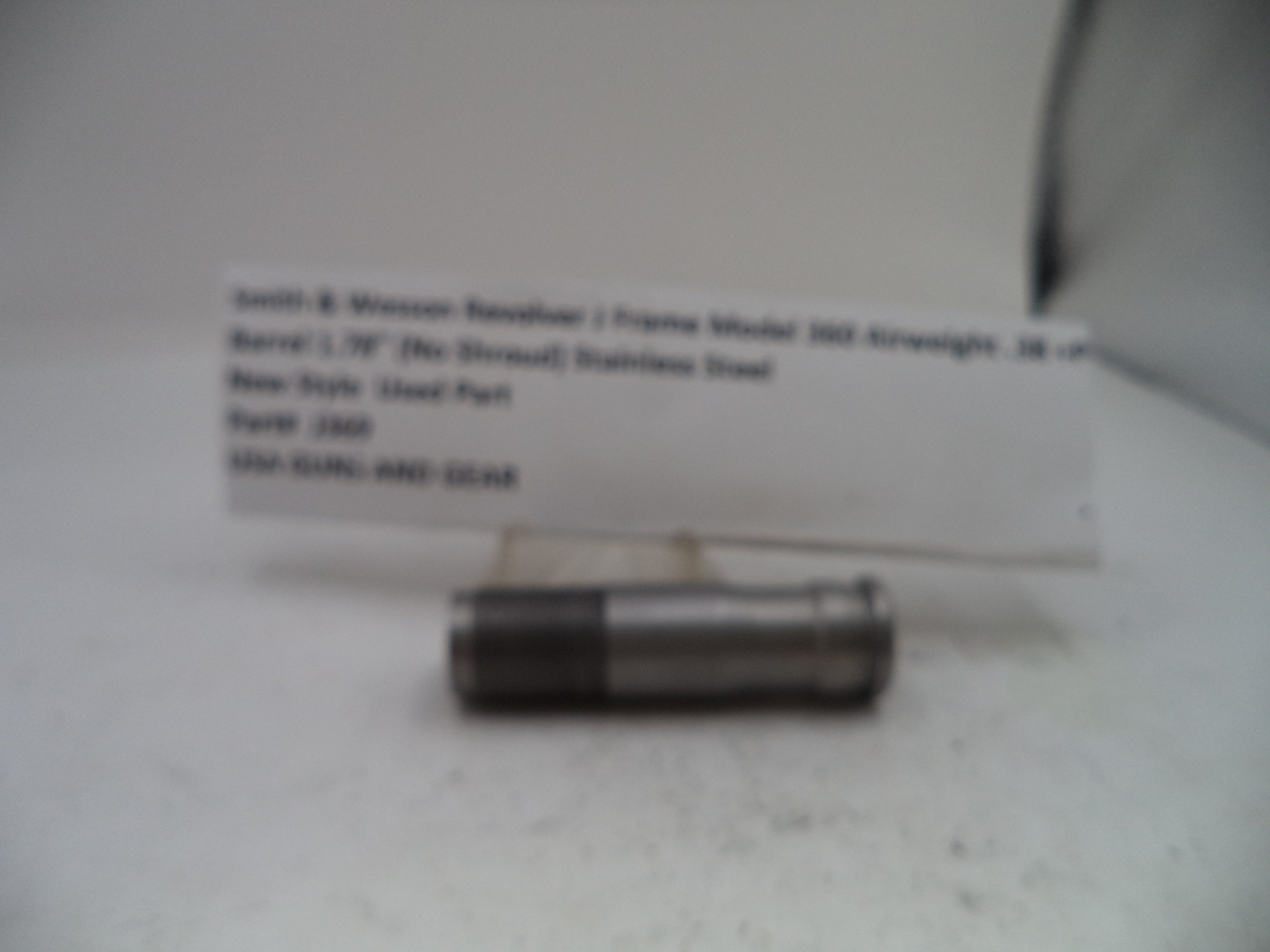 A small cylindrical metal part with a white label card behind it on a light surface. The label appears to read Smith & Wesson Basilore 1 Frame Model 500 Airstream .