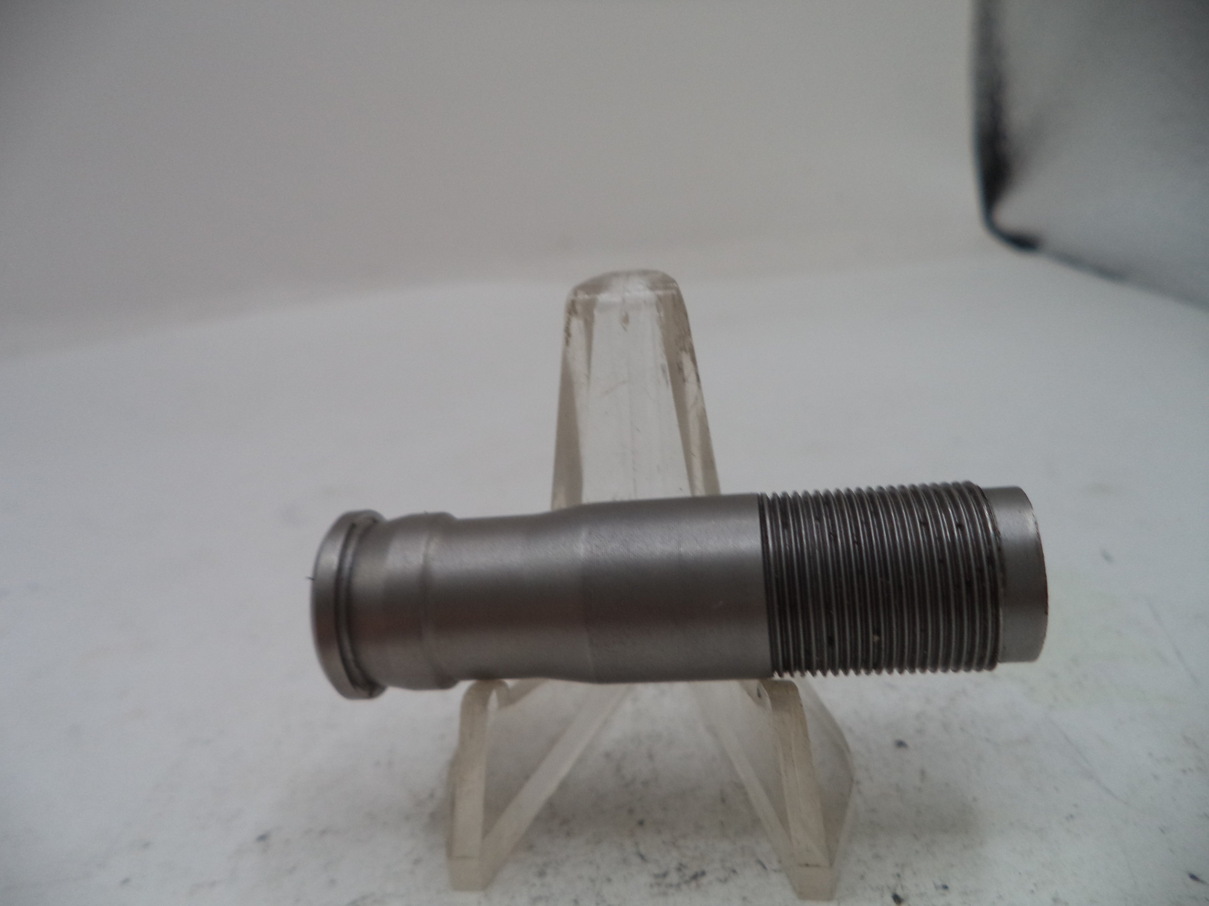 A metallic threaded pipe fitting or coupler shown horizontally on a clear plastic display stand against a plain light background.