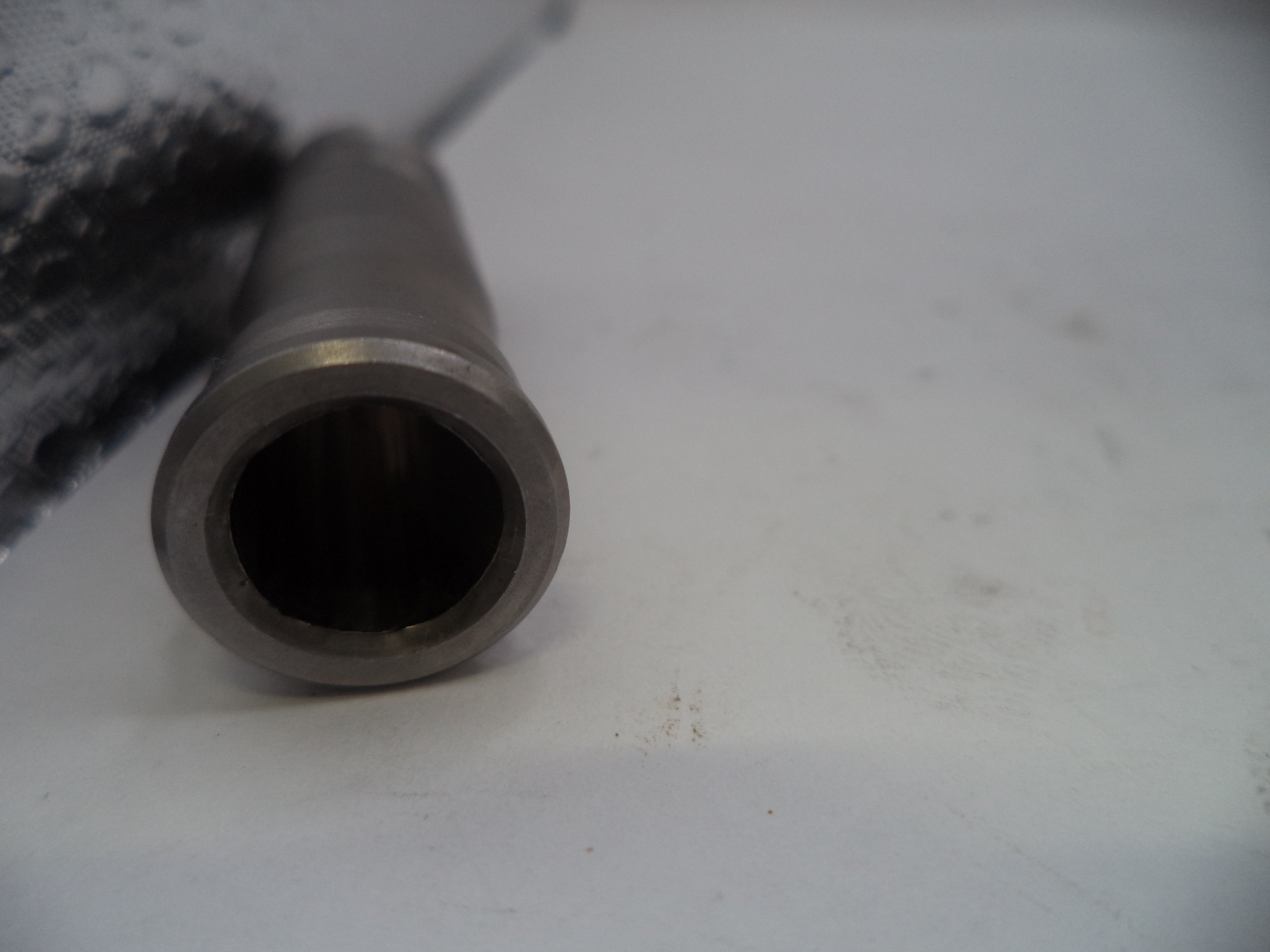 A close-up shot of a metallic cylindrical component, showing a machined silver-gray end with a dark interior, resting on a light gray surface.