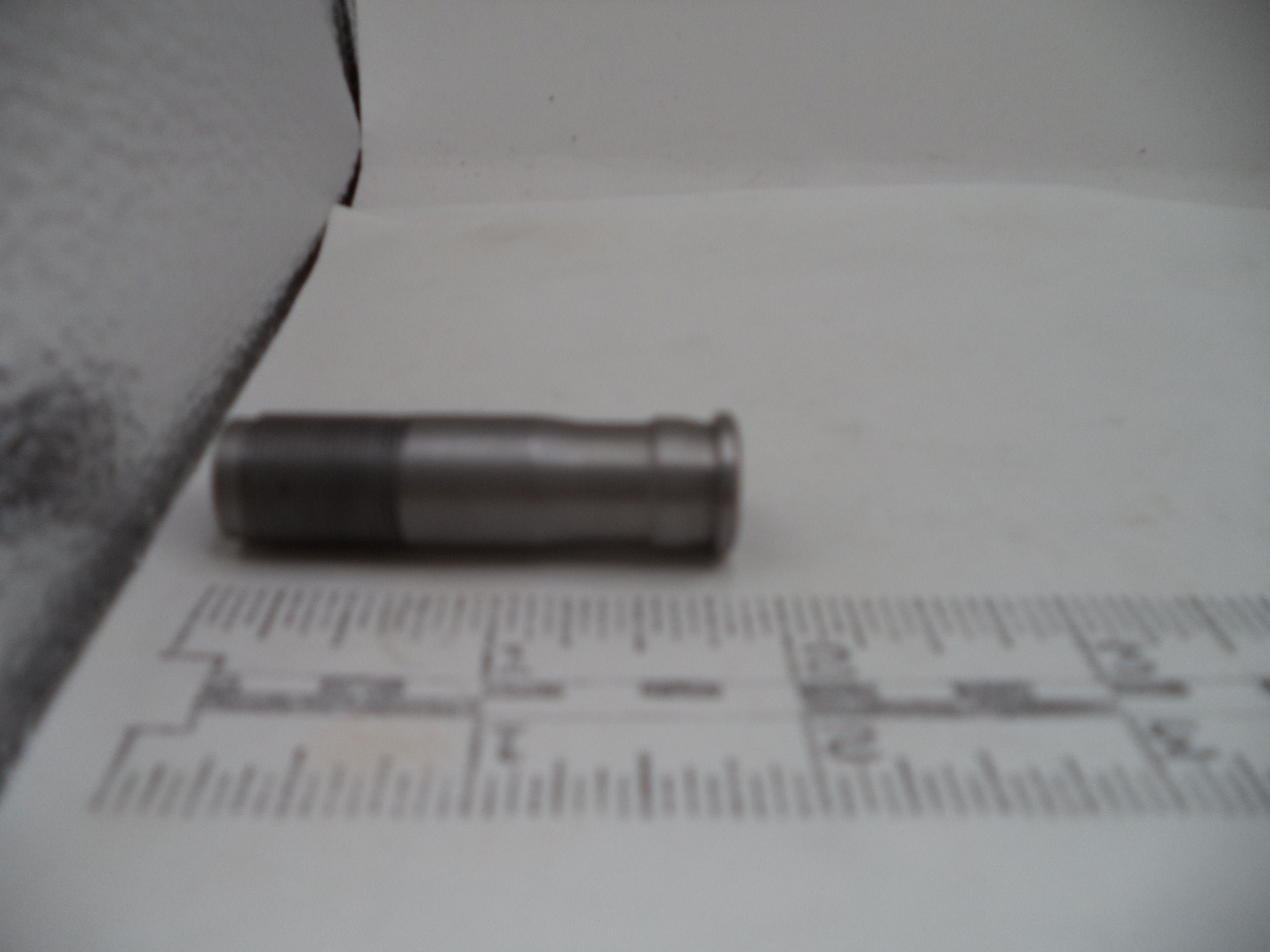A small metallic cylindrical object, possibly a bullet casing or prop, resting on a white surface with a ruler in the foreground for scale.