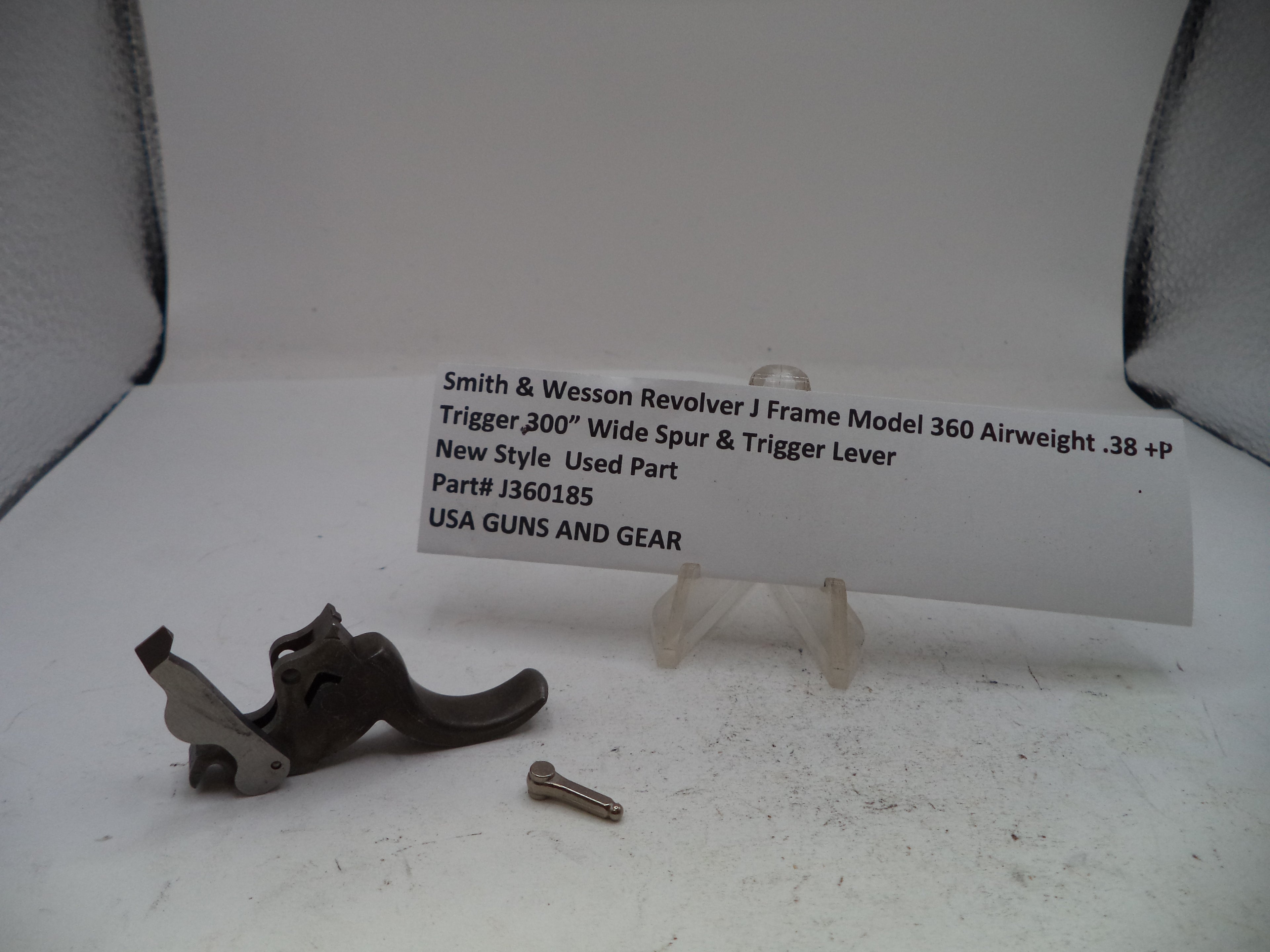 A used Smith & Wesson revolver trigger lever and screw on a white surface, with a white label card displaying part details and the text USA GUNS AND GEAR.