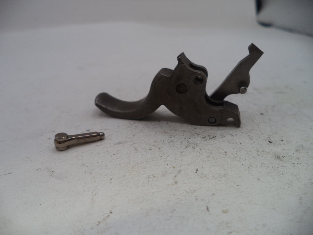 A dark metal firearm trigger assembly with a small matching metal pin or bolt placed beside it on a light gray surface.