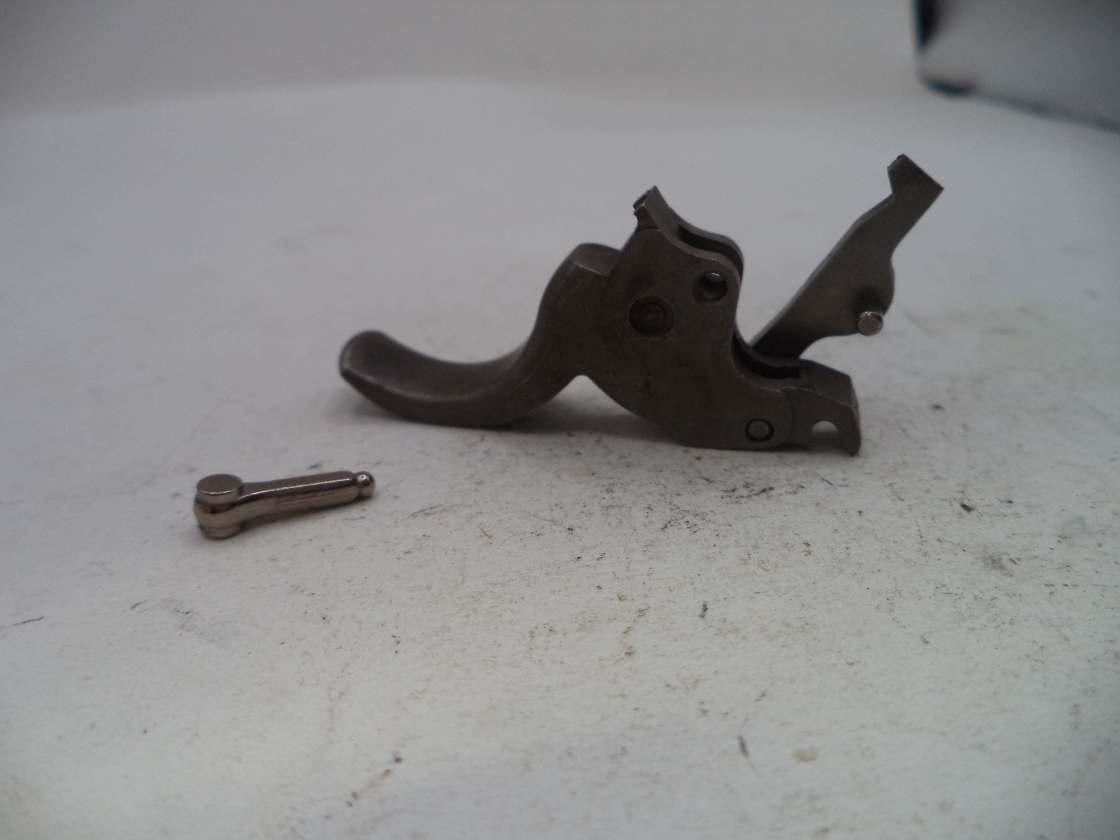 A dark metal firearm trigger assembly with a small matching metal pin or bolt placed beside it on a light gray surface.