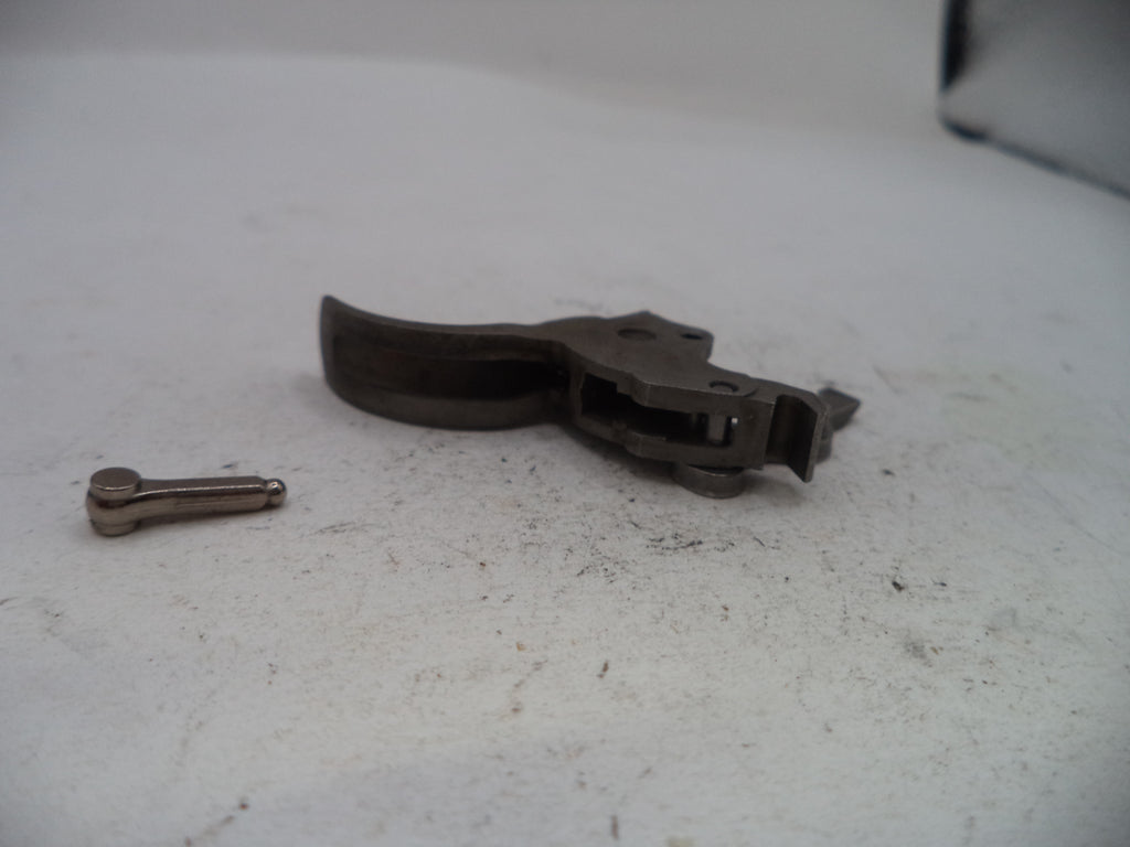 A dark metal trigger assembly and a small metal pin placed on a light gray, slightly dusty surface.