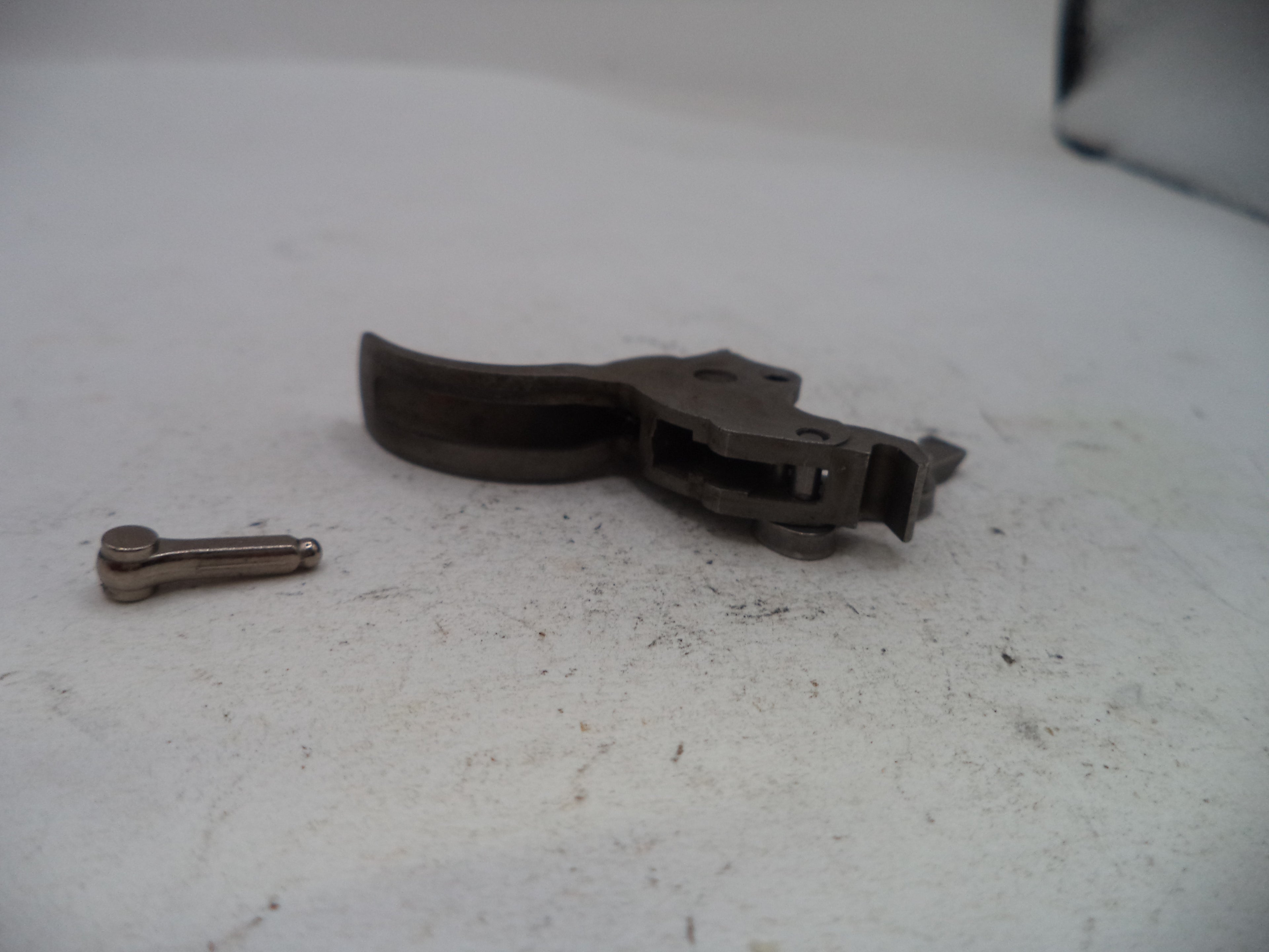 A dark metal trigger assembly and a small metal pin placed on a light gray, slightly dusty surface.