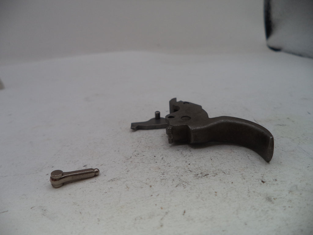 A disassembled metal trigger assembly with a curved dark metal grip and a small separate metal pin, placed on a light gray surface.