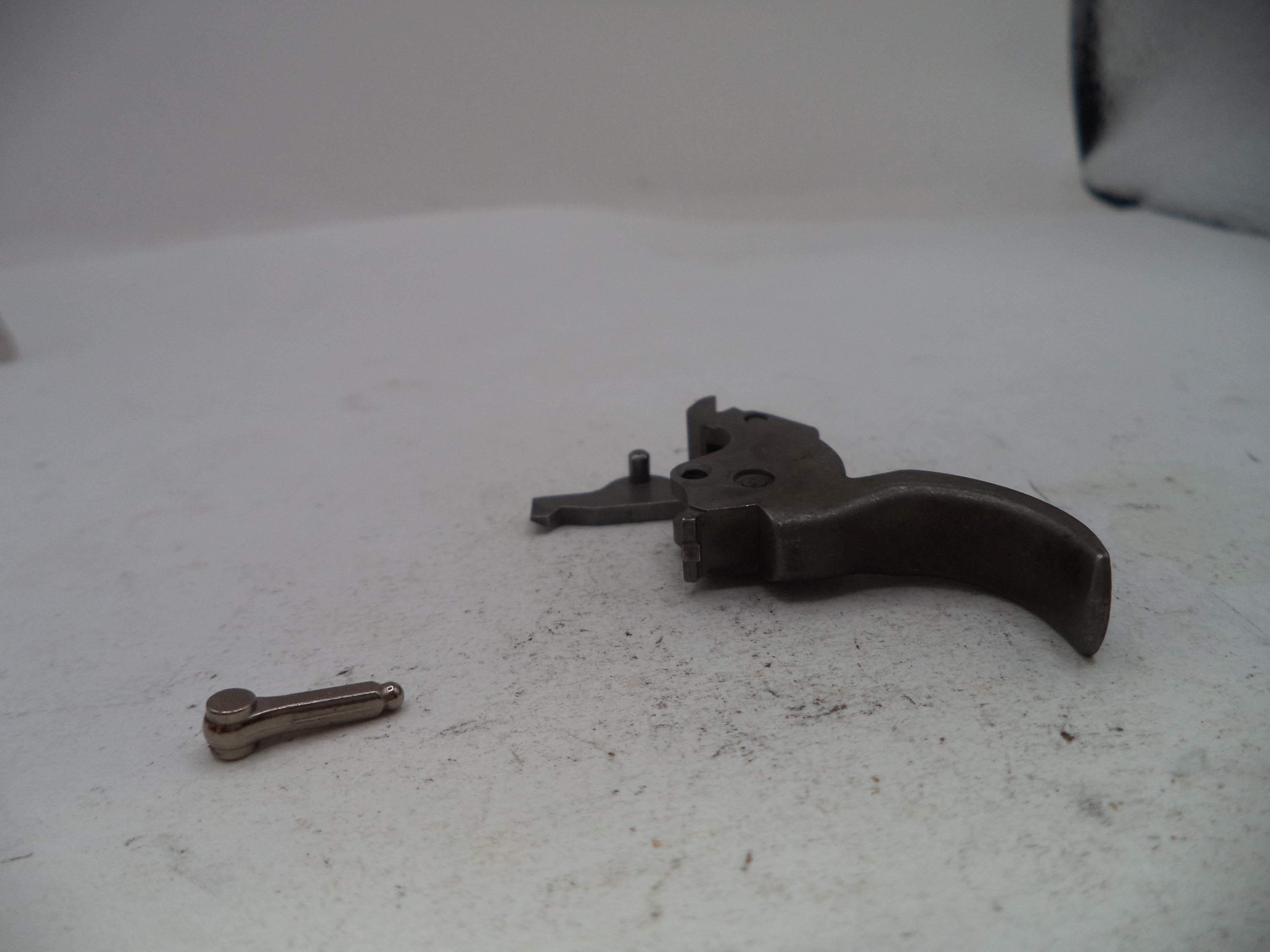 A disassembled metal trigger assembly with a curved dark metal grip and a small separate metal pin, placed on a light gray surface.