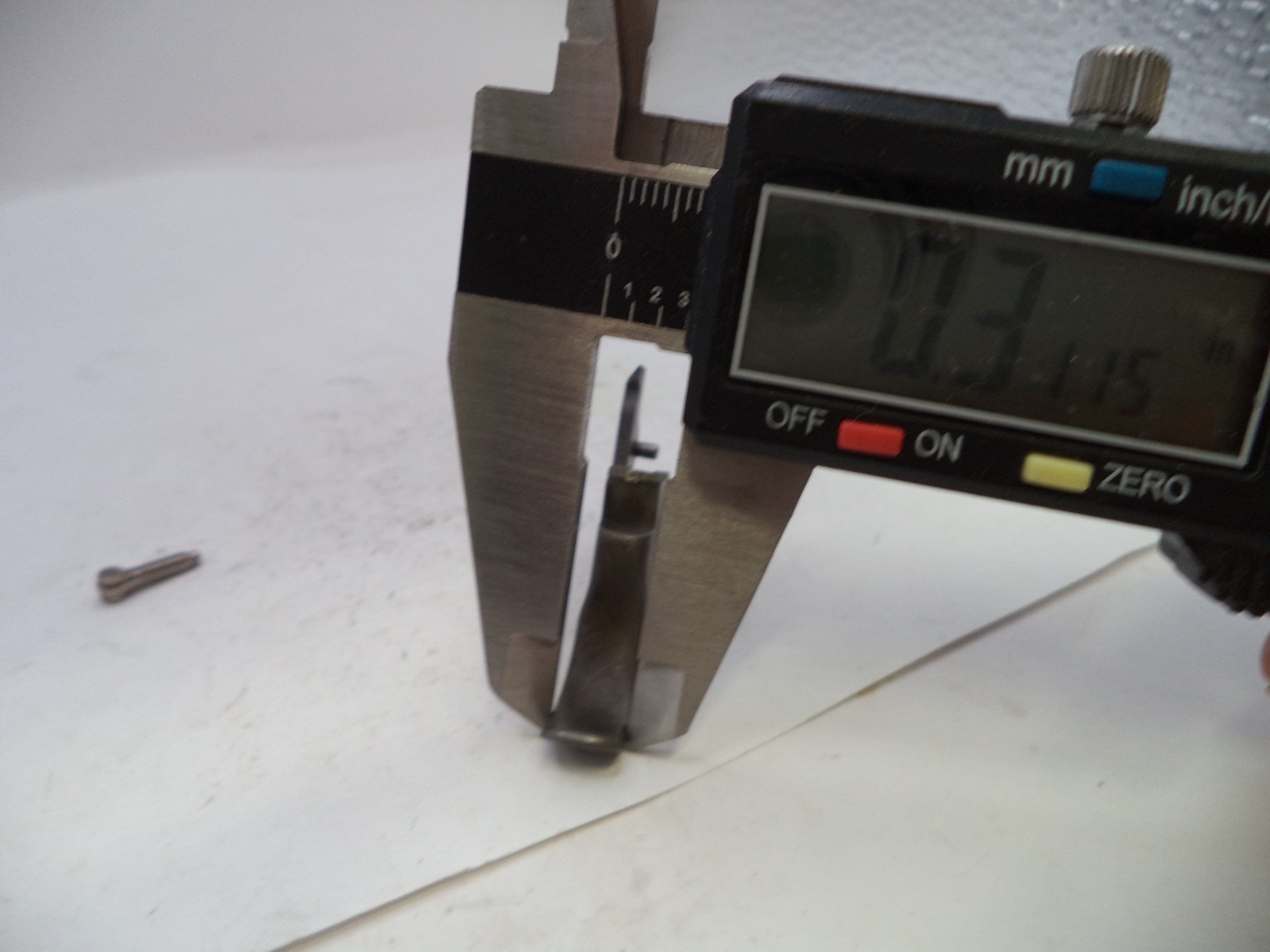 A close-up of a digital caliper measuring a small metal component, with a loose matching screw on a white surface nearby.
