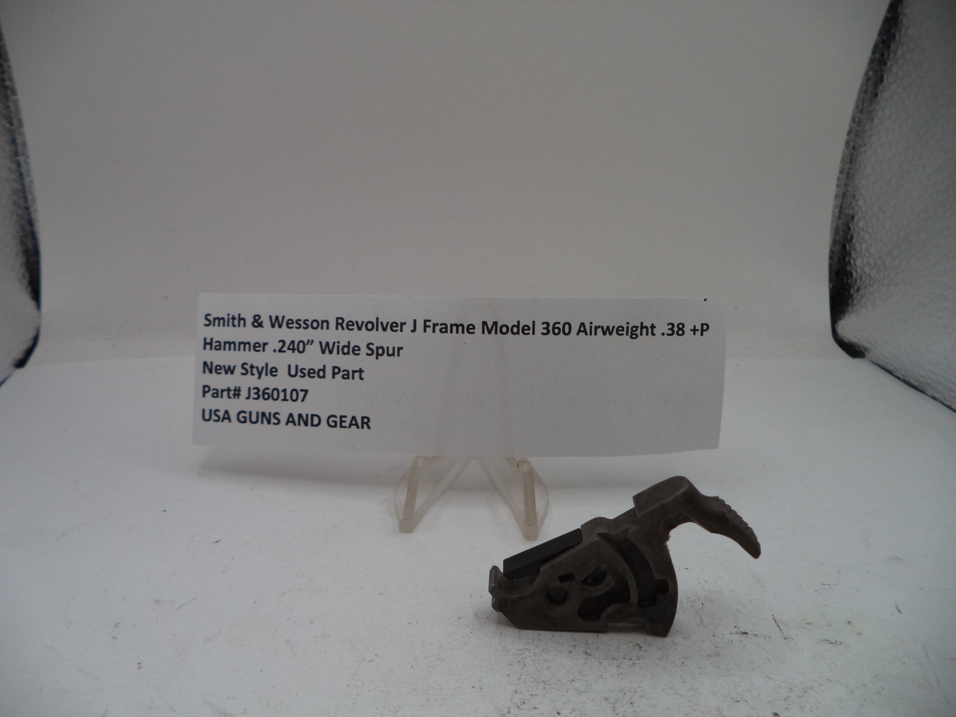 A used metal hammer component for a Smith & Wesson revolver, displayed on a small clear stand next to a white paper label card on a white surface.