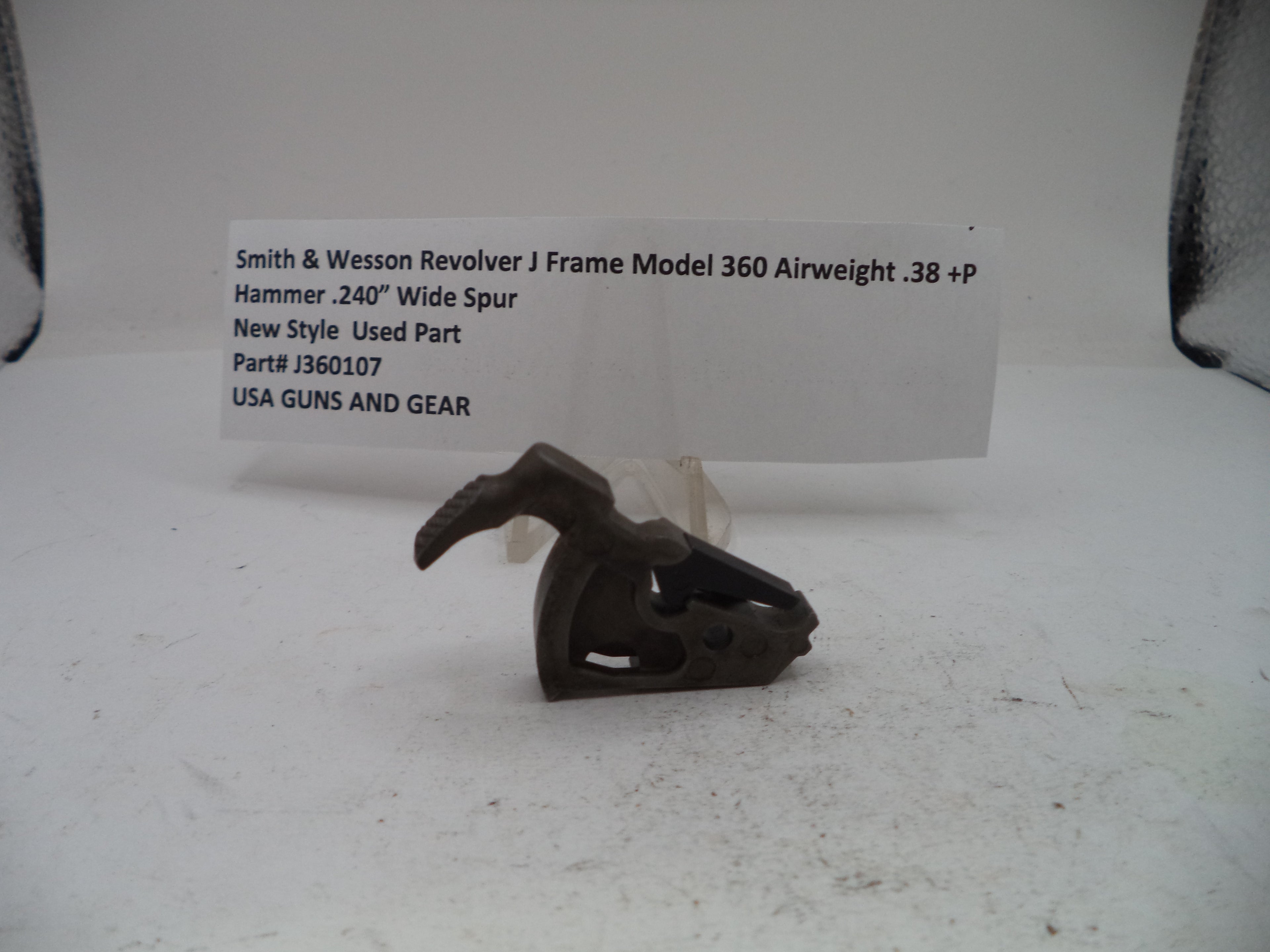 A small used metal firearm part, specifically a Smith & Wesson revolver J frame hammer, displayed on a white surface with a paper label card behind it.