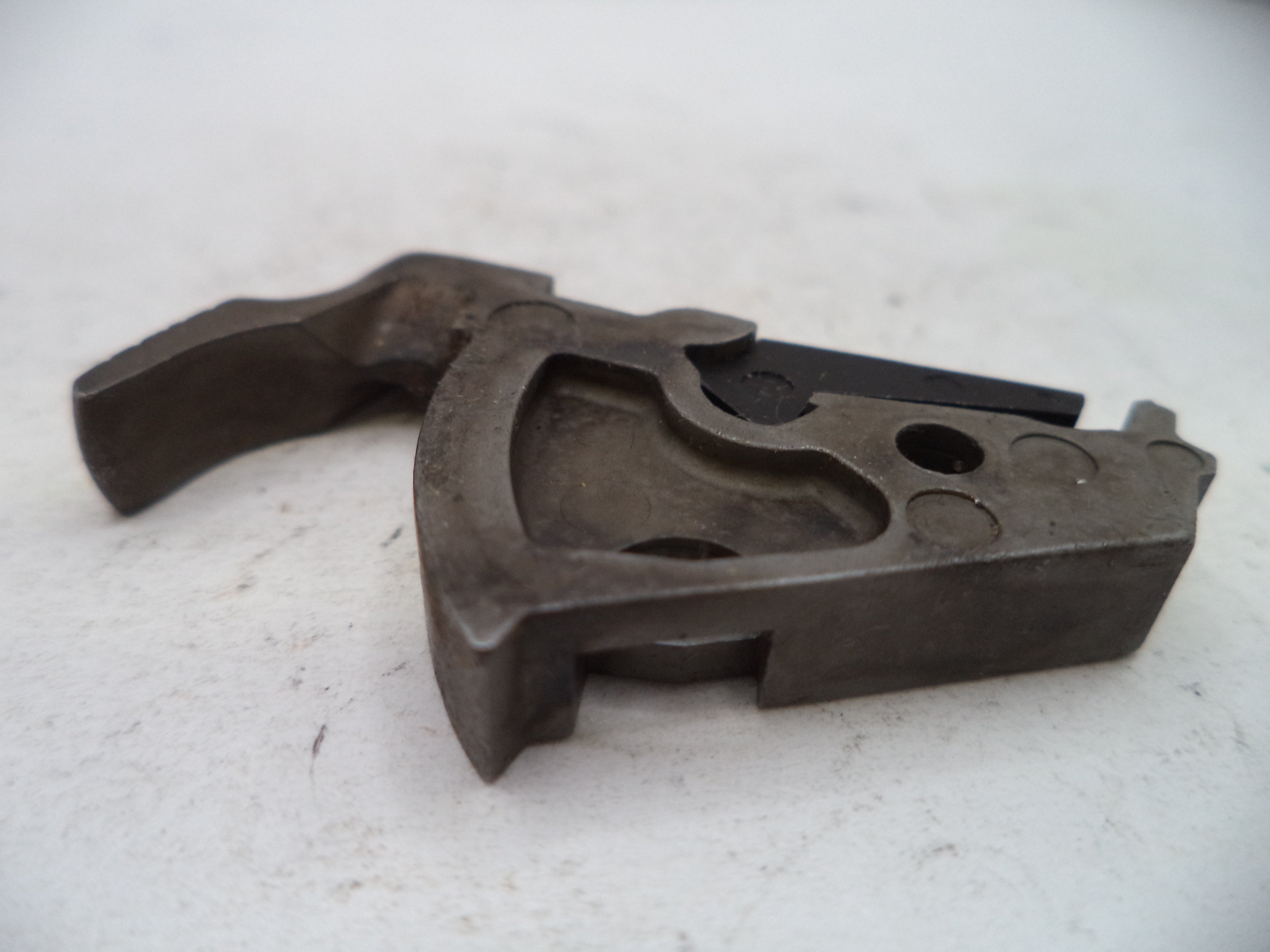 A dark metal mechanical component or tool part with a curved handle and circular cutouts, photographed on a light off-white surface.