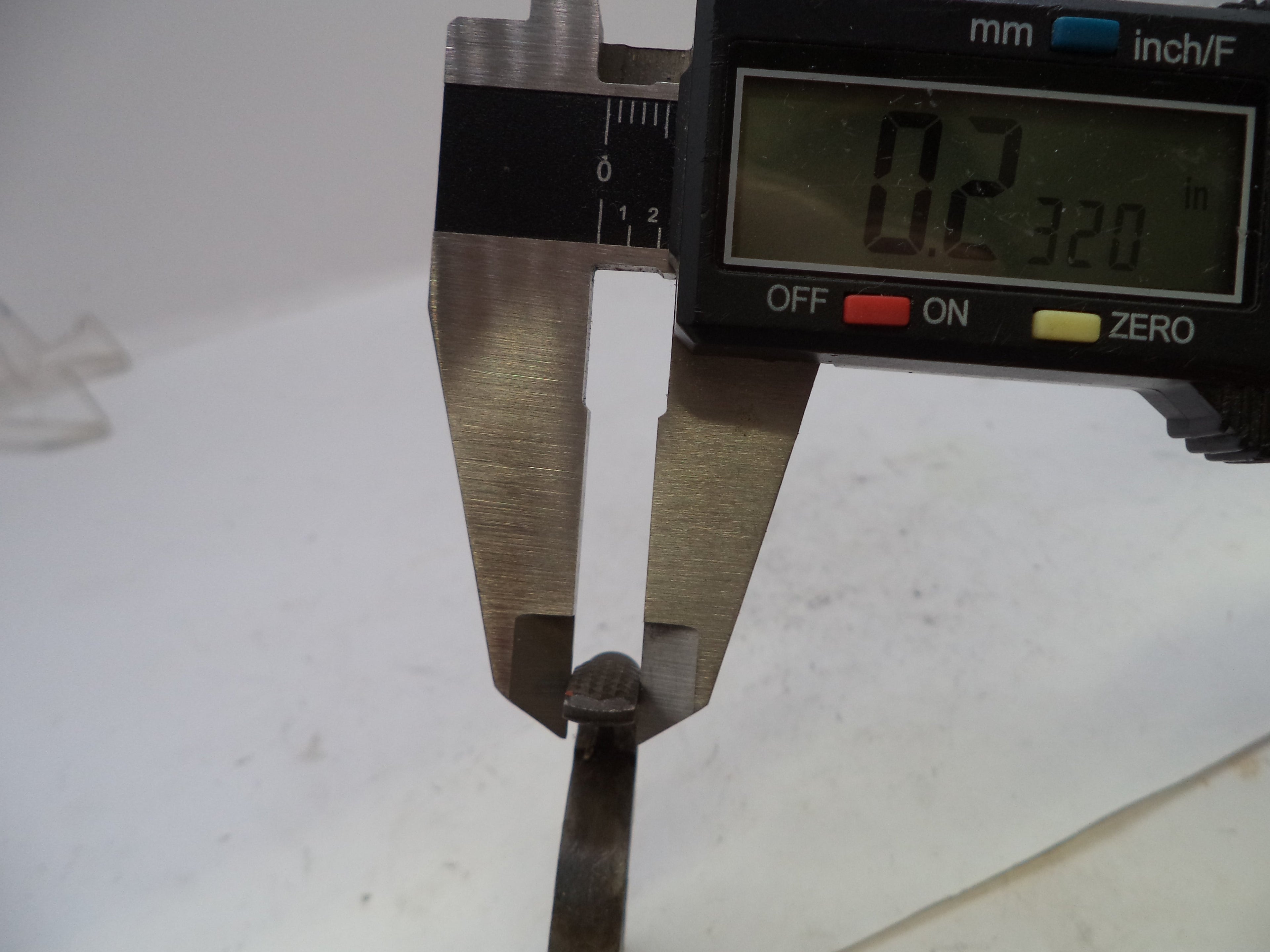 Digital caliper measuring the thickness of a small metal shim or gasket on a white surface.