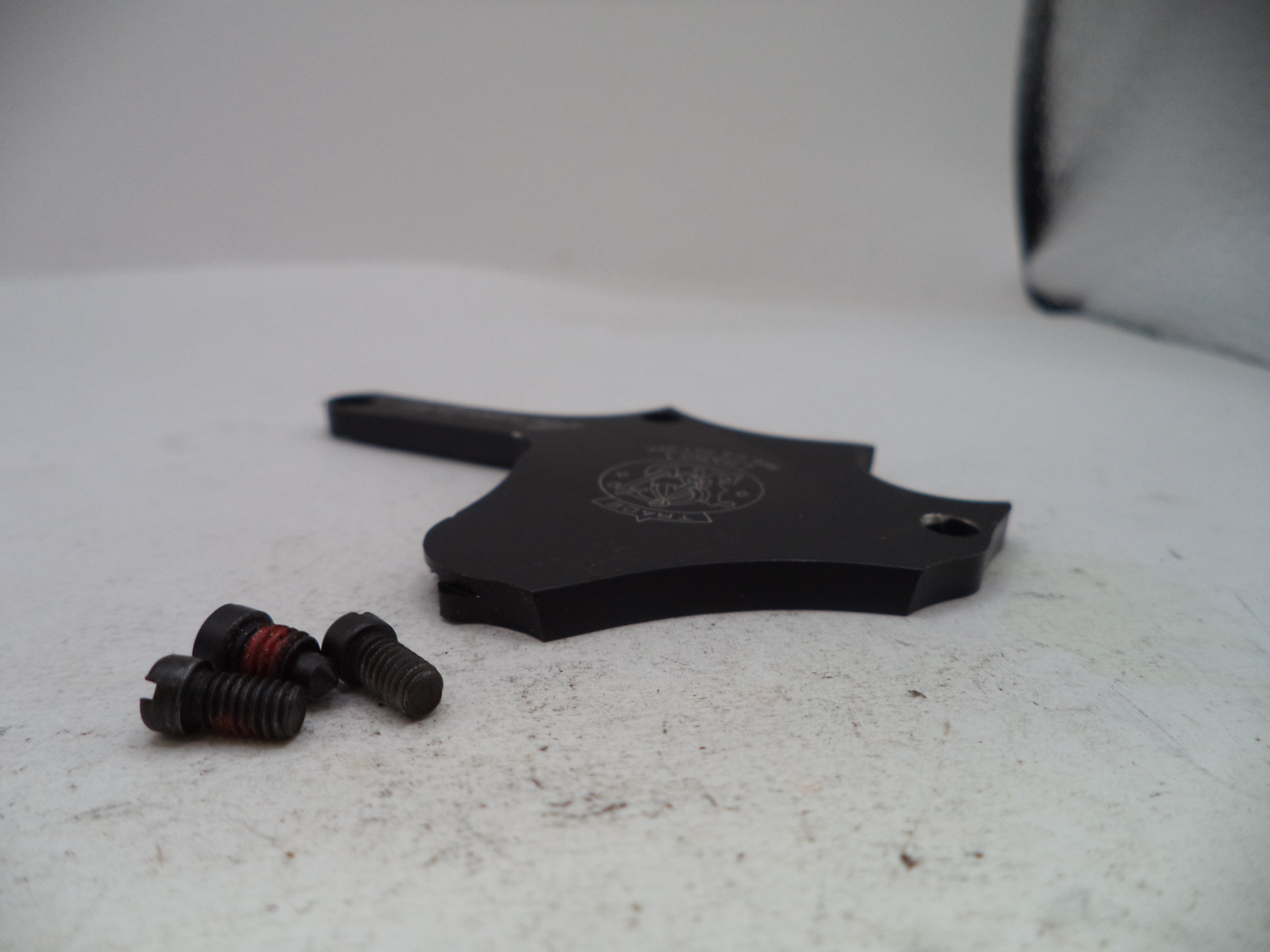 A black metal mounting bracket with a hexagonal profile and three small black screws with red thread wraps, placed on a light gray textured surface.