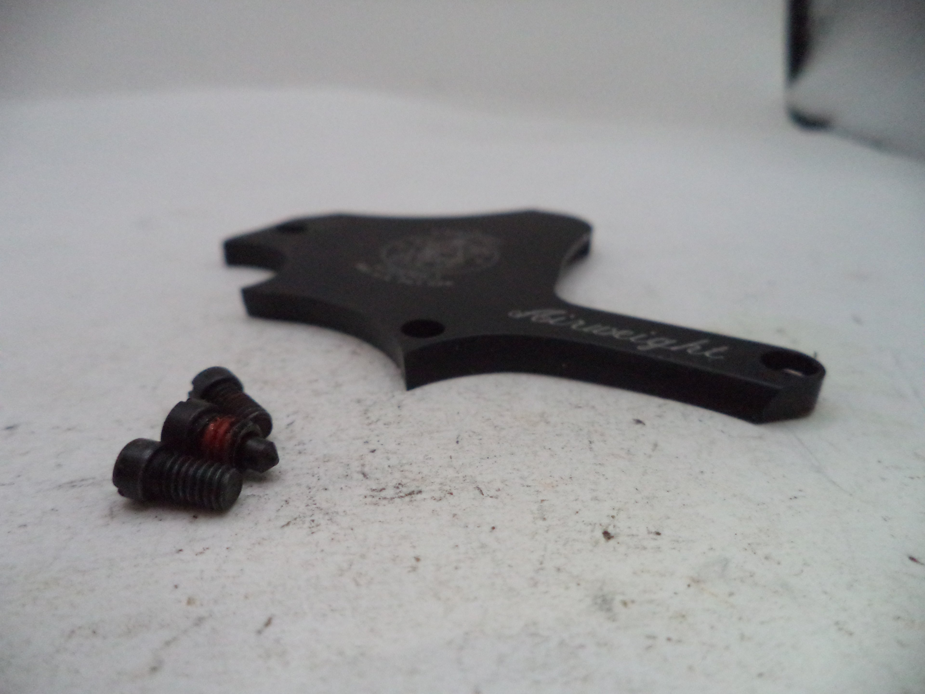 A black metal weight adjustment piece with small threaded adjustment screws, photographed on a light off-white surface.